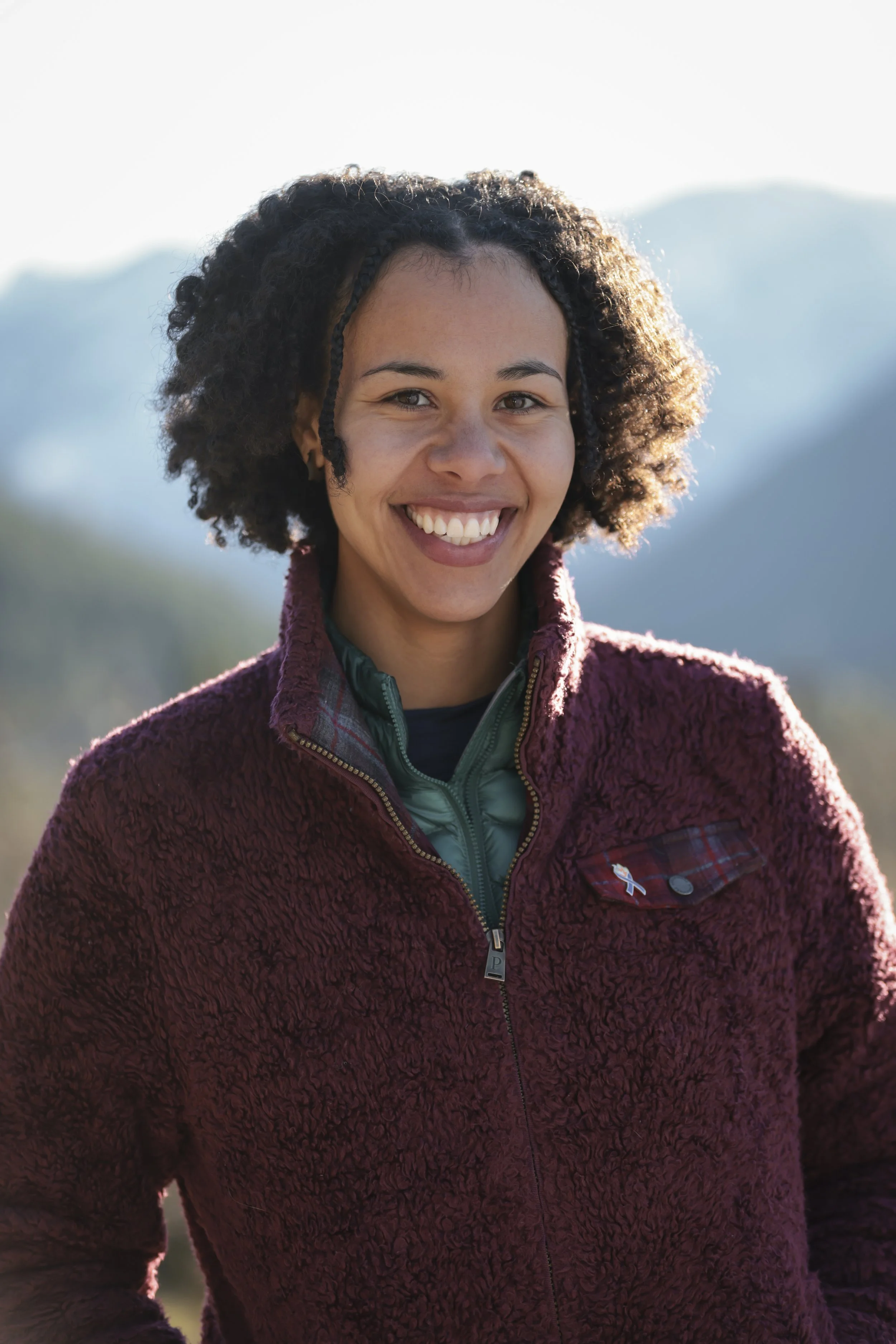 A smiling woman with curly hair wearing a maroon fleece jacket outdoors with mountains in the background.