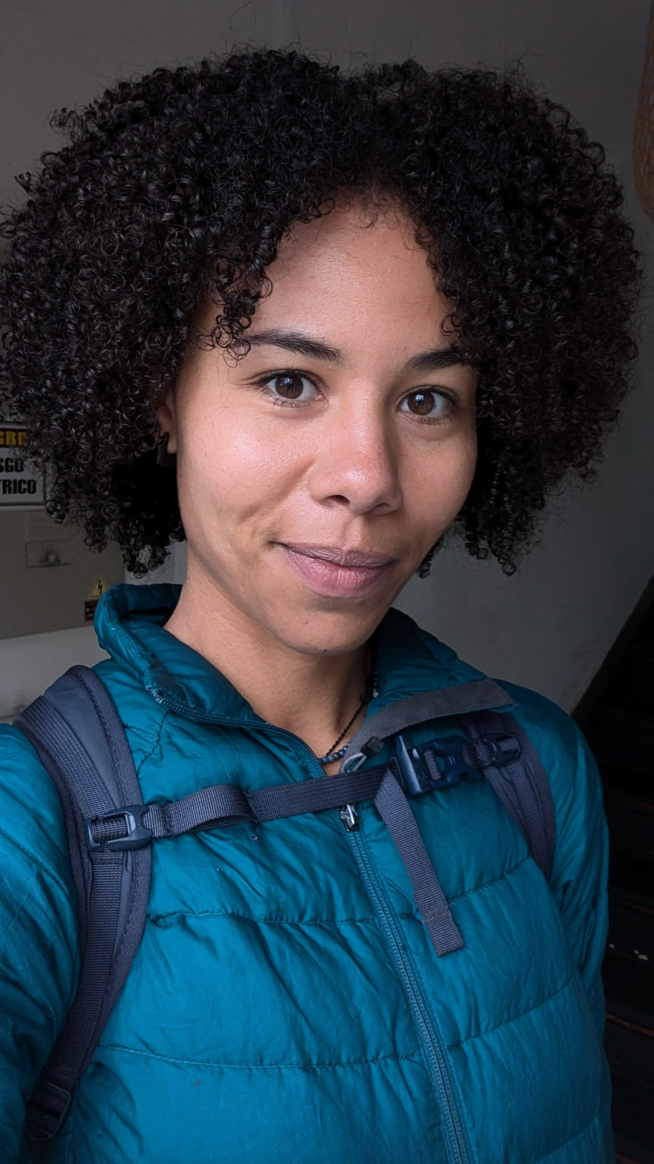 A woman with curly black hair, wearing a teal puffer jacket and a backpack, taking a selfie indoors with a neutral expression.