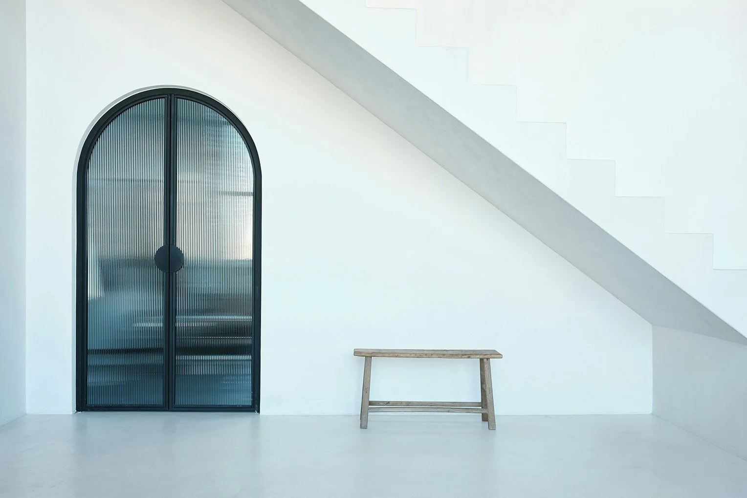 A bare, modern minimalistic space with white walls and floor. A single bench sits in the space next to arching reeded glass french doors under a stairway.