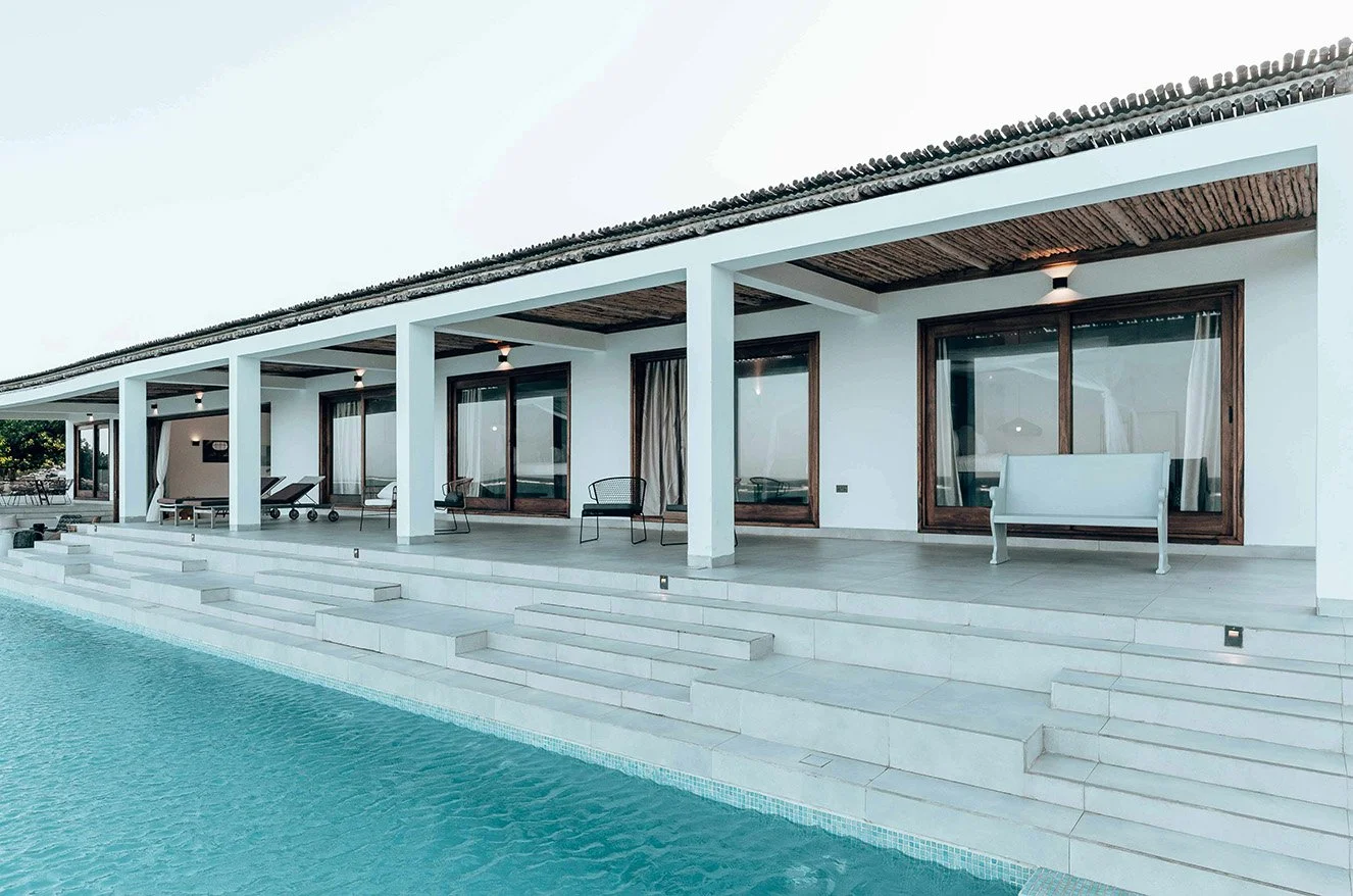A row of resort bungalows. Each bungalow has a large wood-framed glass sliding door and they are slightly elevated above a large pool.