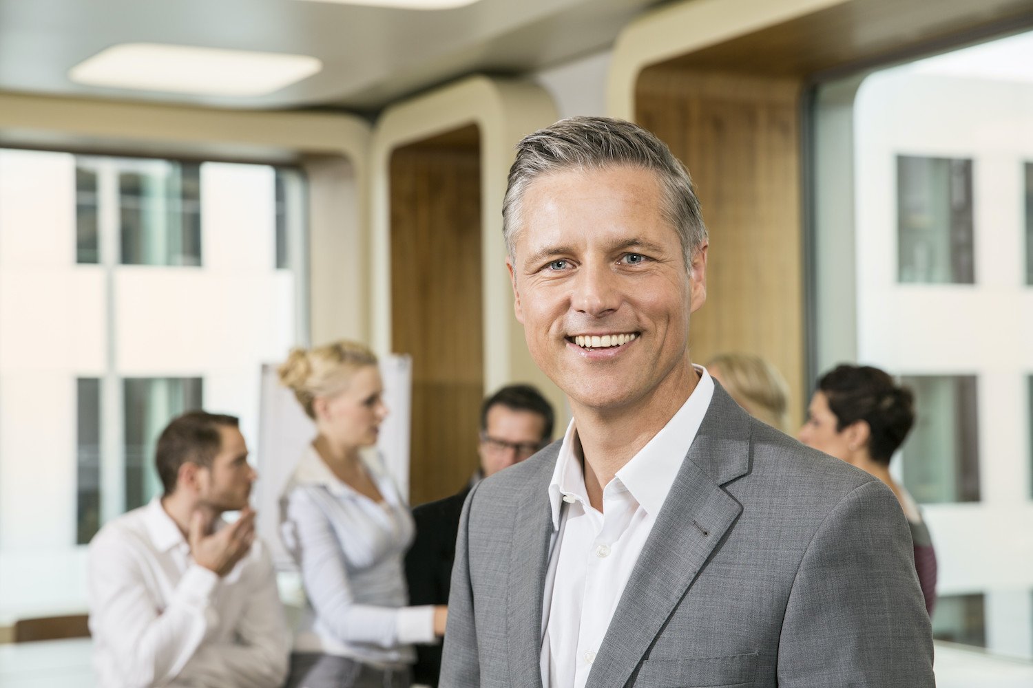 Smiling man in a gray suit at a business meeting or networking event, with a group of people in the background engaged in conversation.