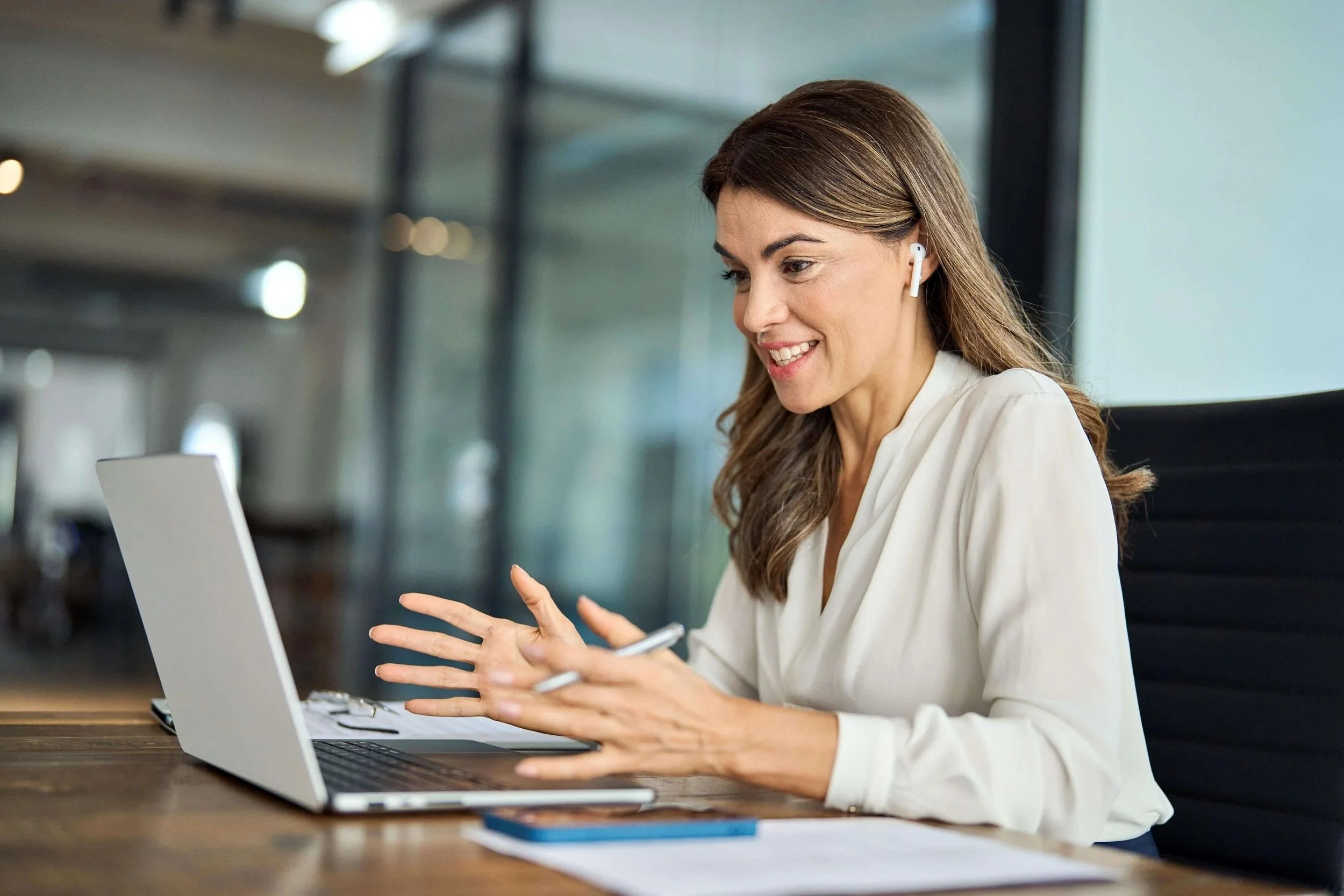 A woman sitting at a wooden desk with a laptop, wearing wireless earphones and smiling while holding a pen.