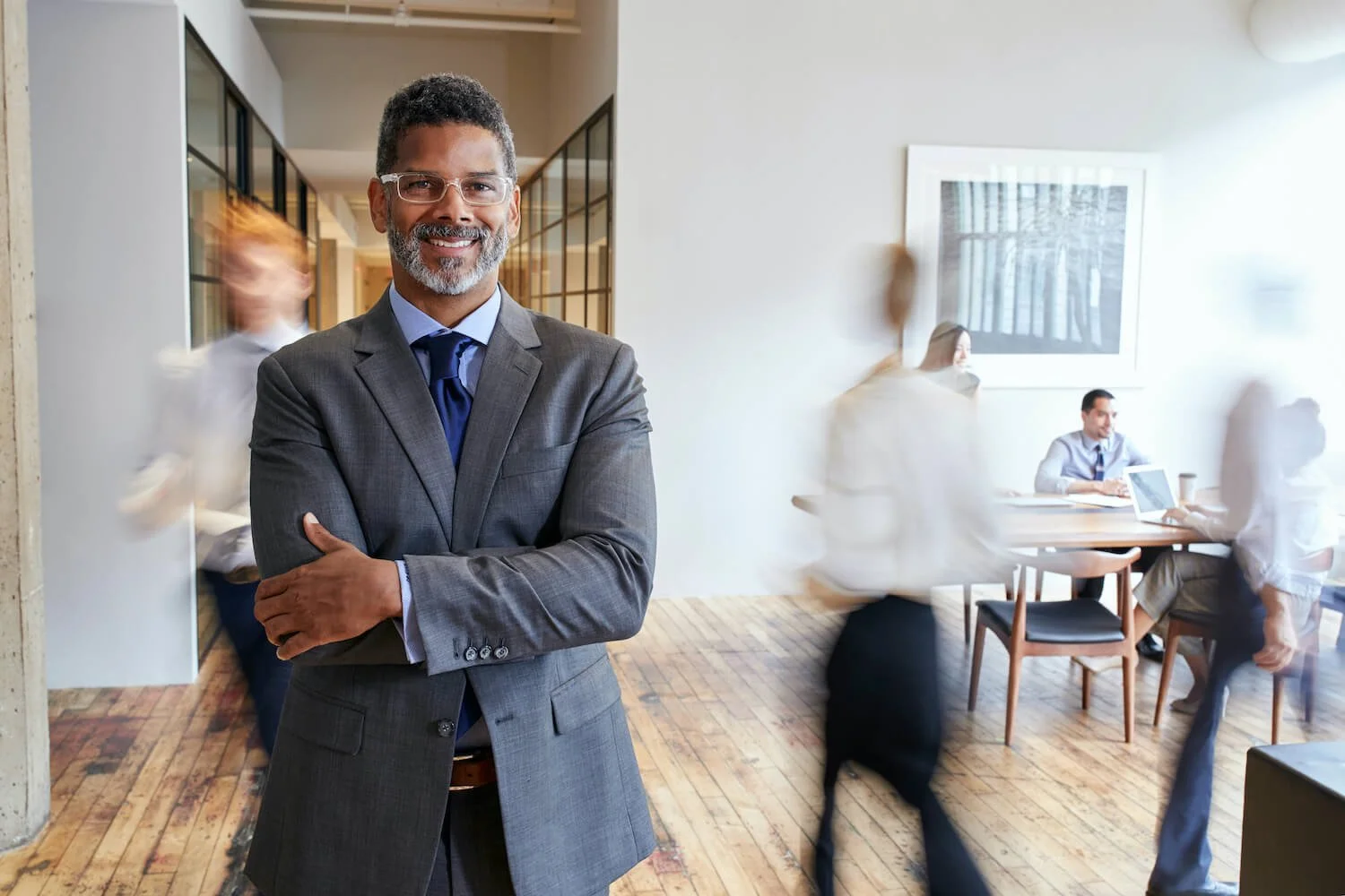 A confident middle-aged man in a gray suit with glasses smiling and standing with arms crossed in an office or conference room with people working and walking in the background.