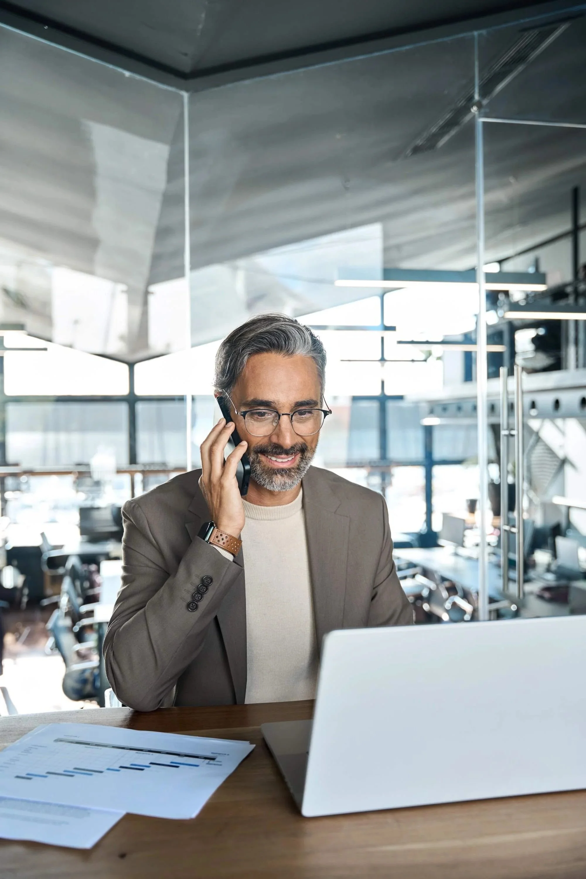 A middle-aged man with glasses, a beard, and gray hair is talking on his cell phone while sitting at a desk with documents and a laptop in front of him. He is wearing a beige sweater and a brown blazer. The background shows a modern office space with large windows, desks, and office equipment.