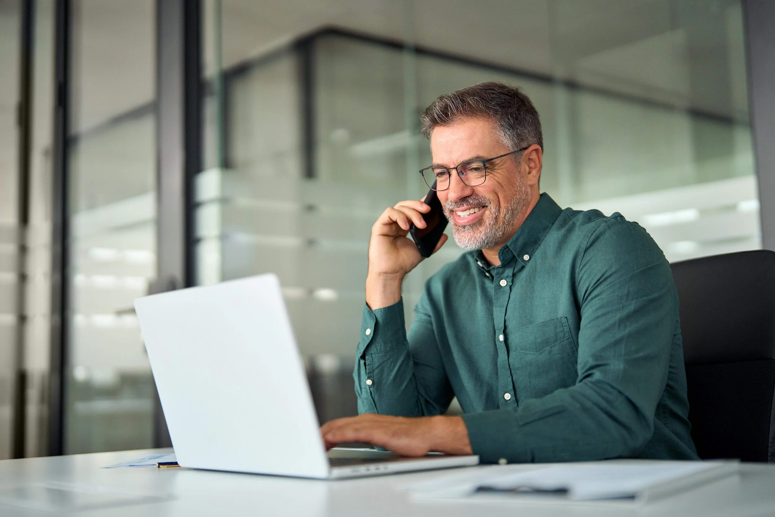 A middle-aged man with glasses and a beard, wearing a green button-up shirt, smiling while talking on his cellphone in an office setting, with a laptop open in front of him.