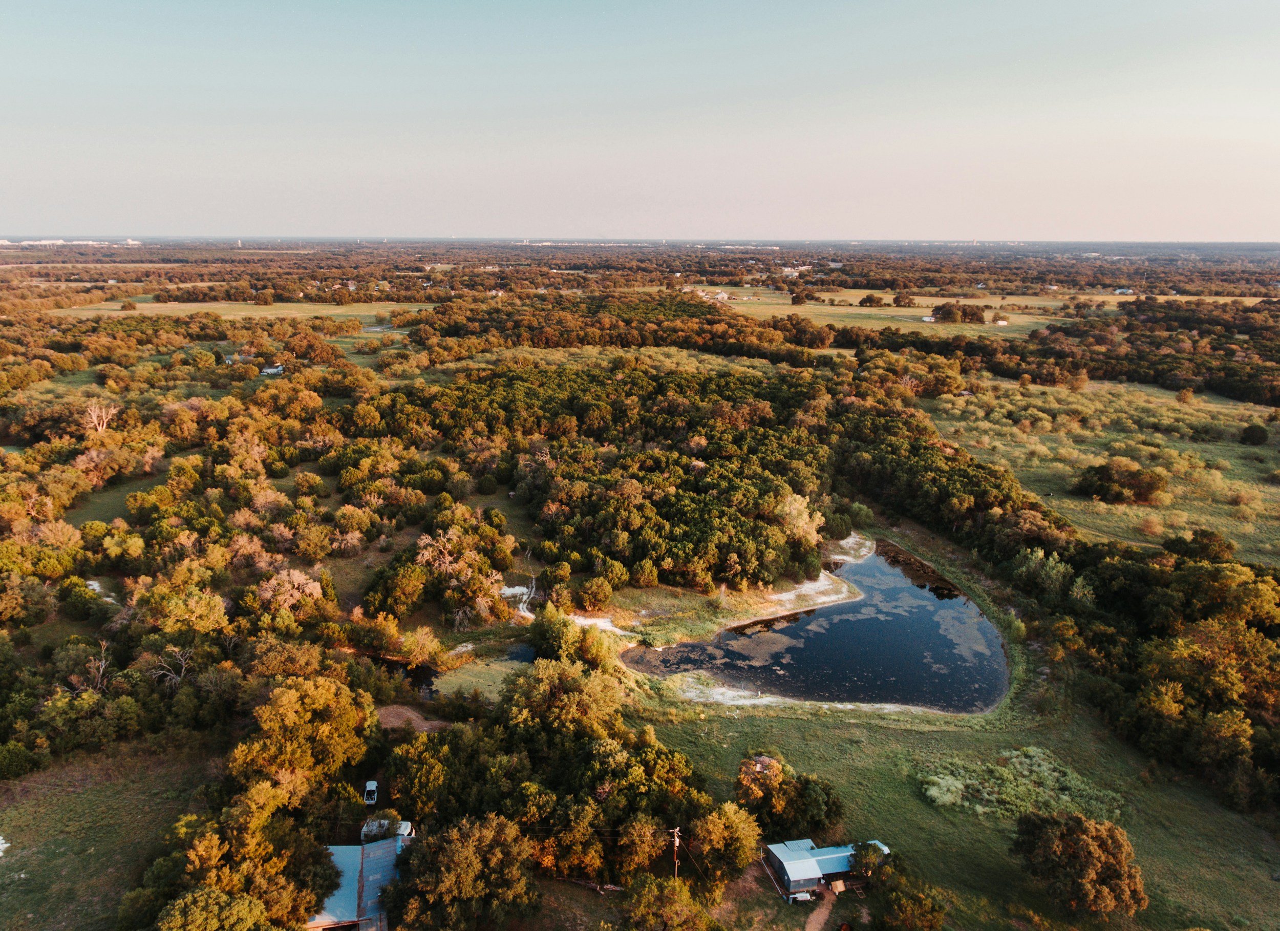Aerial view of a rural landscape with a pond, surrounded by trees and fields, under a clear sky.