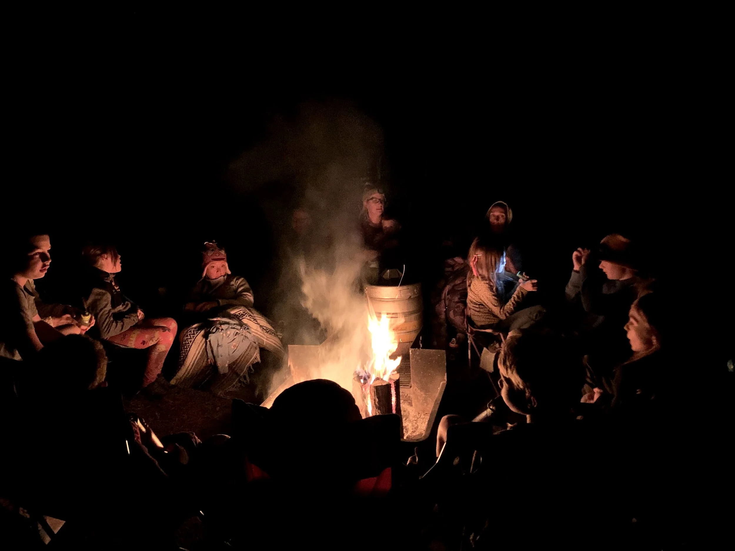 Group of people gathered around a campfire at night, sitting on benches or chairs, some wearing hoodies and hats, with some looking into the fire and others talking. The scene is dark with firelight illuminating their faces.
