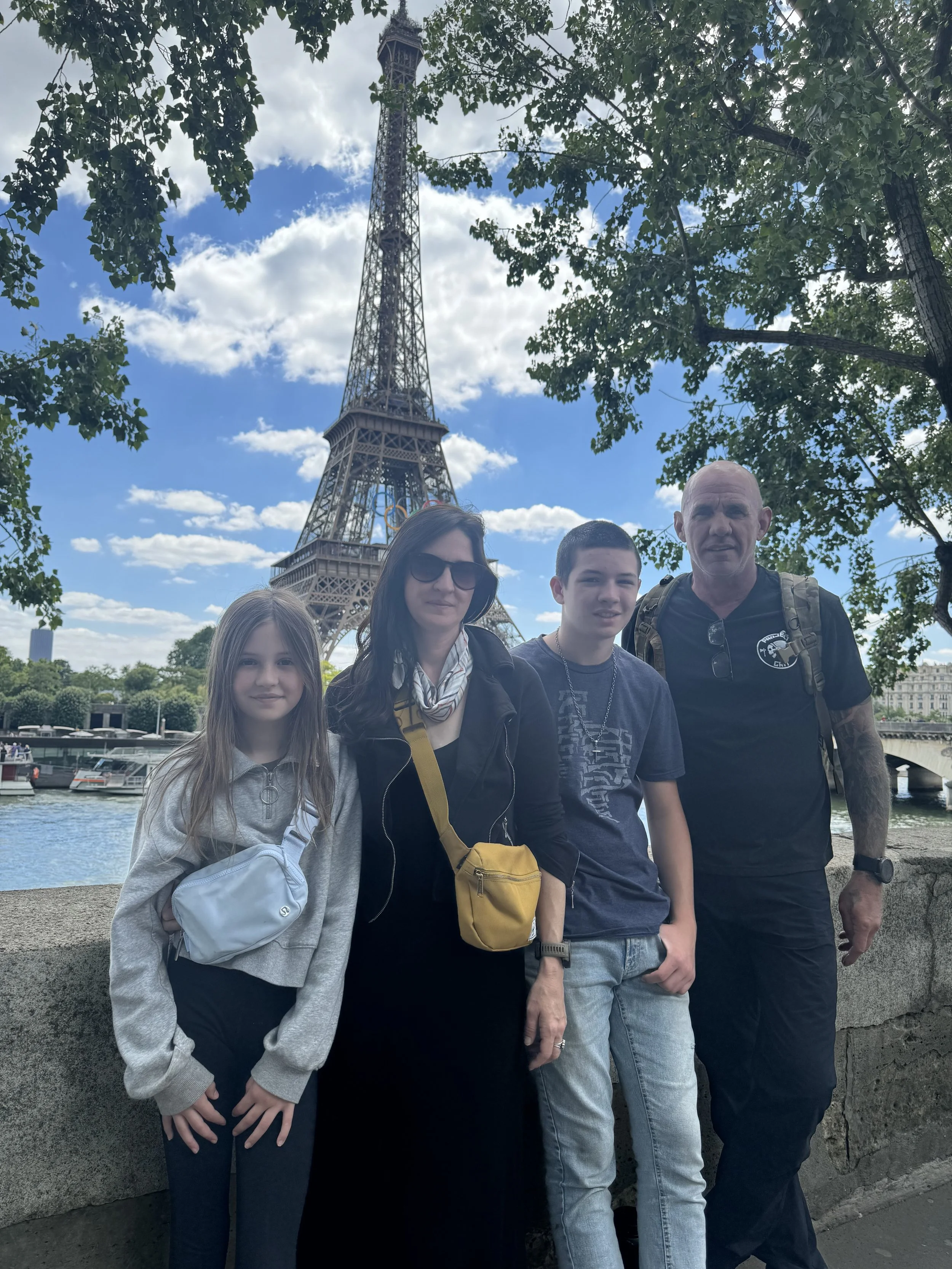 A family of four posing in front of the Eiffel Tower in Paris, France. The group includes a girl, a woman, a teenage boy, and a man, standing by the river with boats and trees surrounding them.