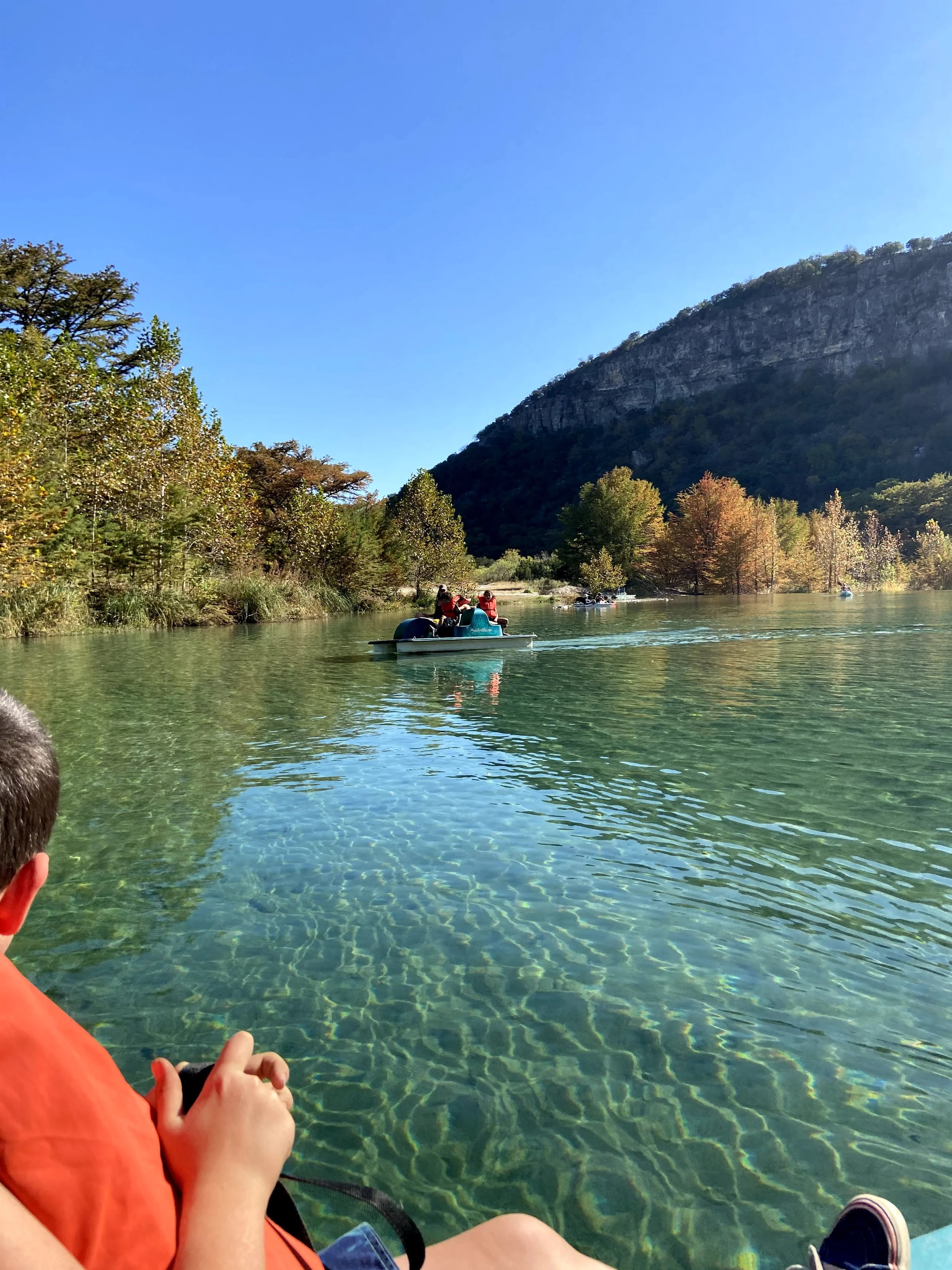 People kayaking and enjoying a clear, calm river surrounded by colorful trees, a mountain in the background, and a bright blue sky.