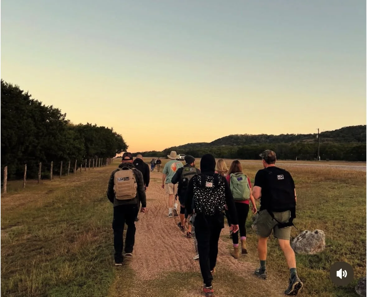 A group of people walking along a dirt trail in an open field during sunset, with some wearing backpacks and hats, and a line of trees on the left side.