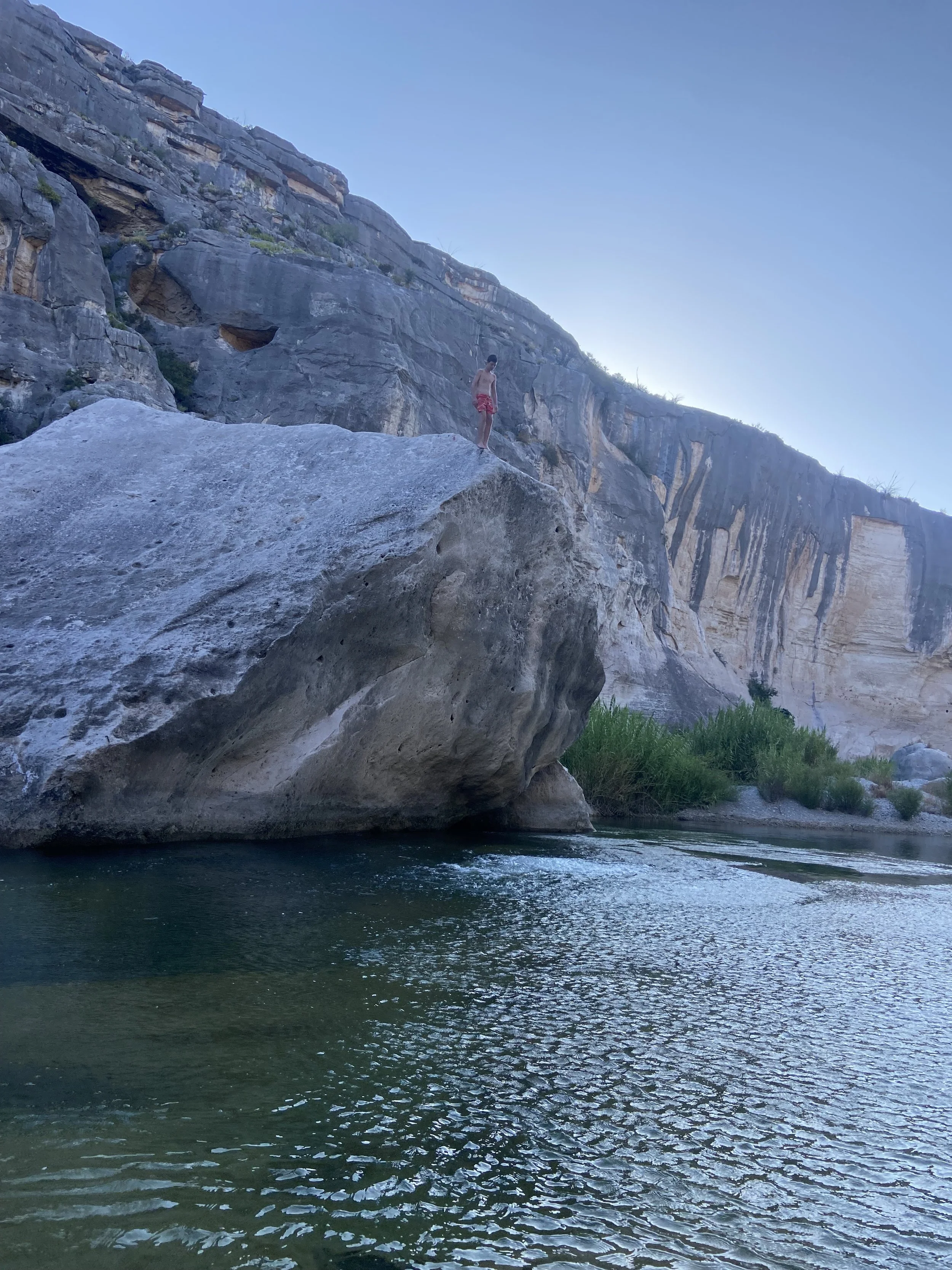 A person wearing red shorts standing on a large rock over water in a canyon with steep rocky cliffs and sparse vegetation, under a clear blue sky.