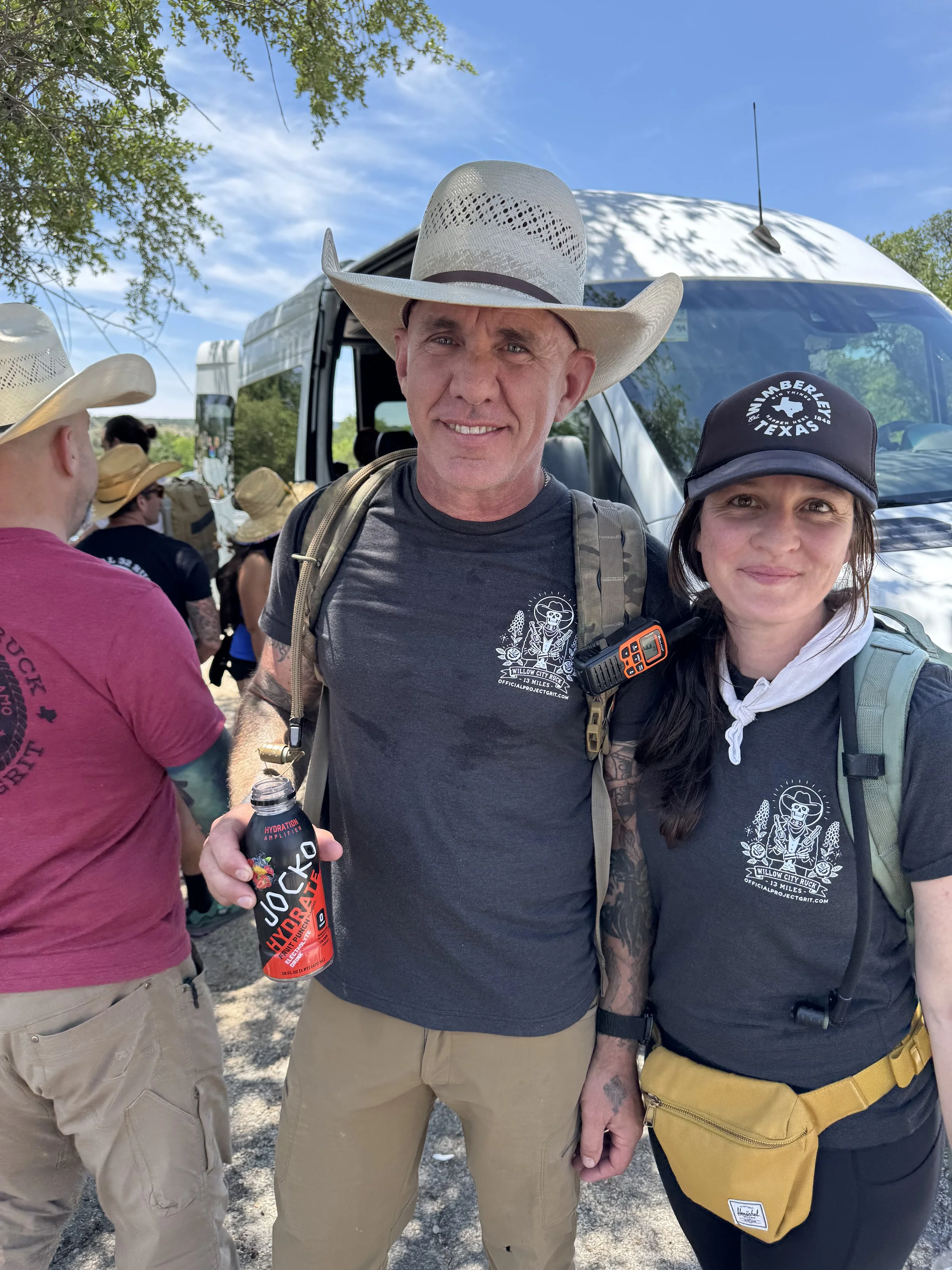 A group of people outdoors, wearing cowboy hats and casual clothing, standing in front of a bus on a sunny day with blue sky and green trees in the background. Two individuals are prominently in the foreground, smiling at the camera.