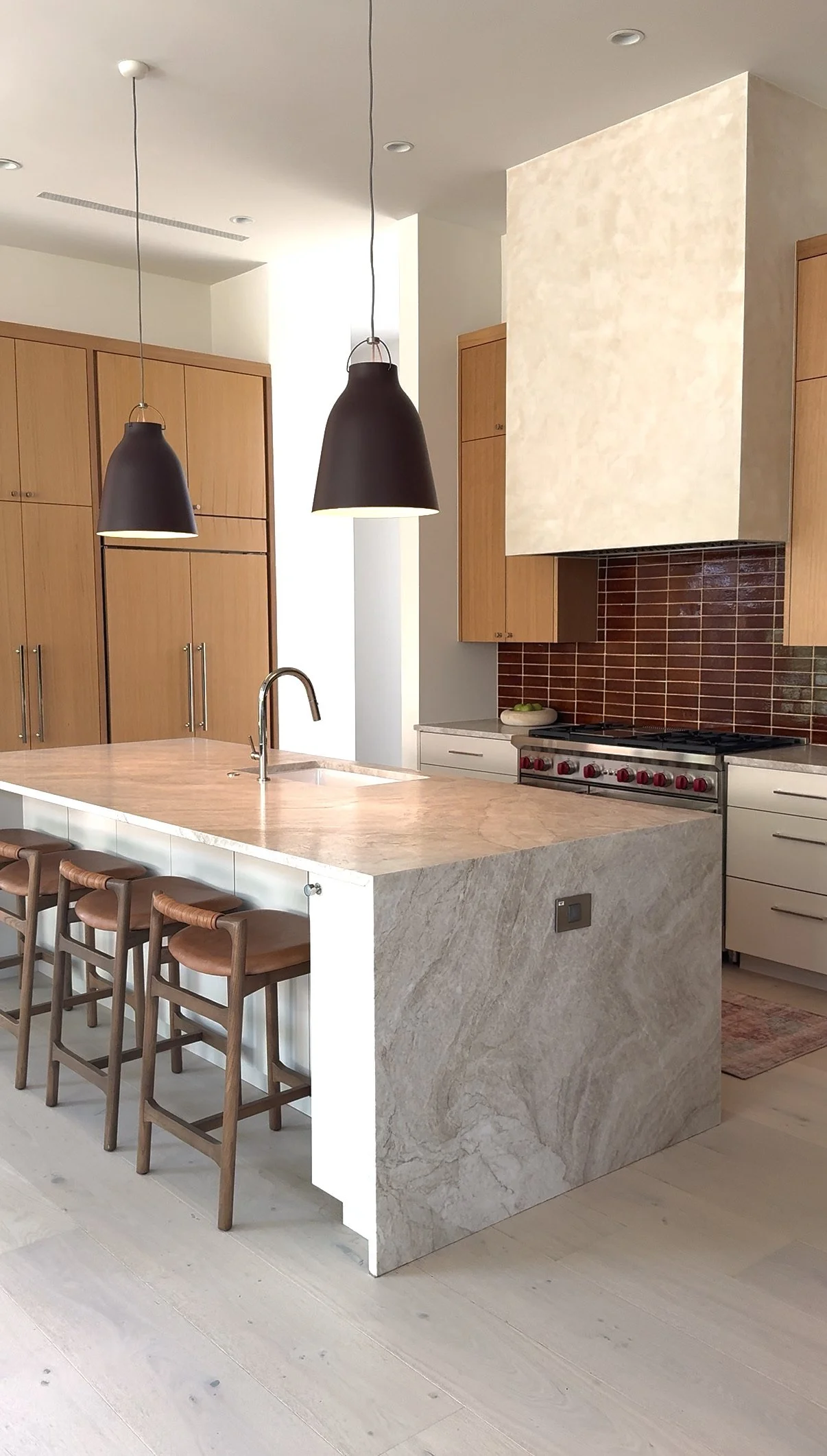 Modern kitchen with beige marble island and brown wooden bar stools. Black pendant lights hanging from the ceiling, light wooden cabinets, and a stove with a brown tile backsplash.