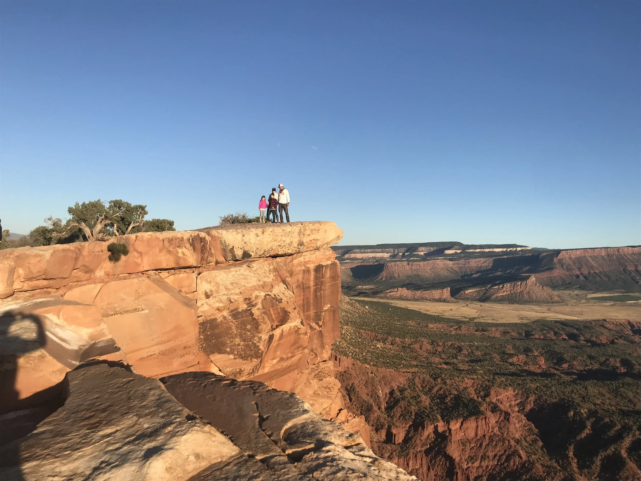 Group of four people standing on the edge of a cliff overlooking a canyon at sunset.