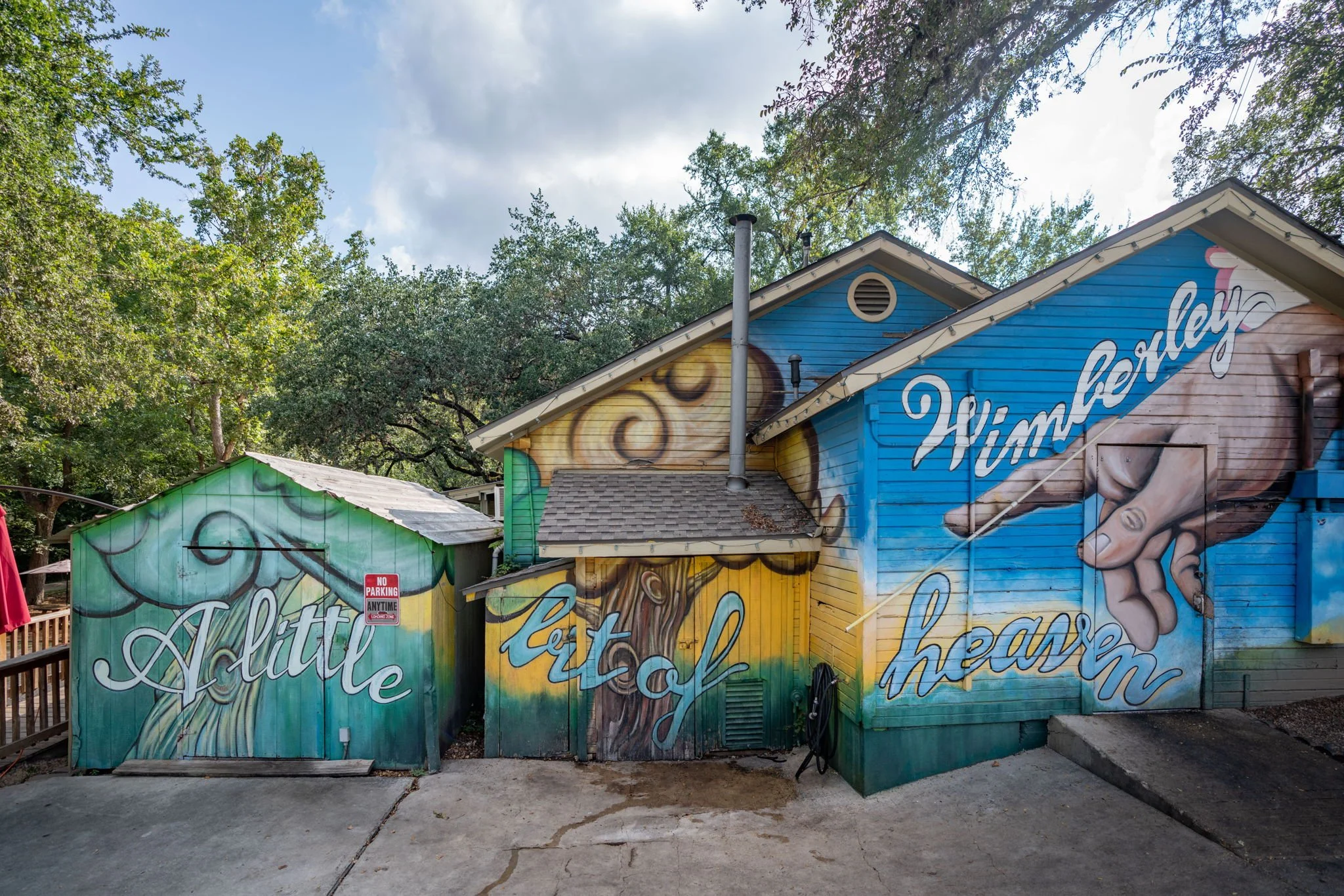 Colorful mural on the side of small buildings depicting a hand holding a baby's hand, with words 'A little' and 'let of' and 'Wimberley' and 'heart' in decorative script, surrounded by trees and cloudy sky.