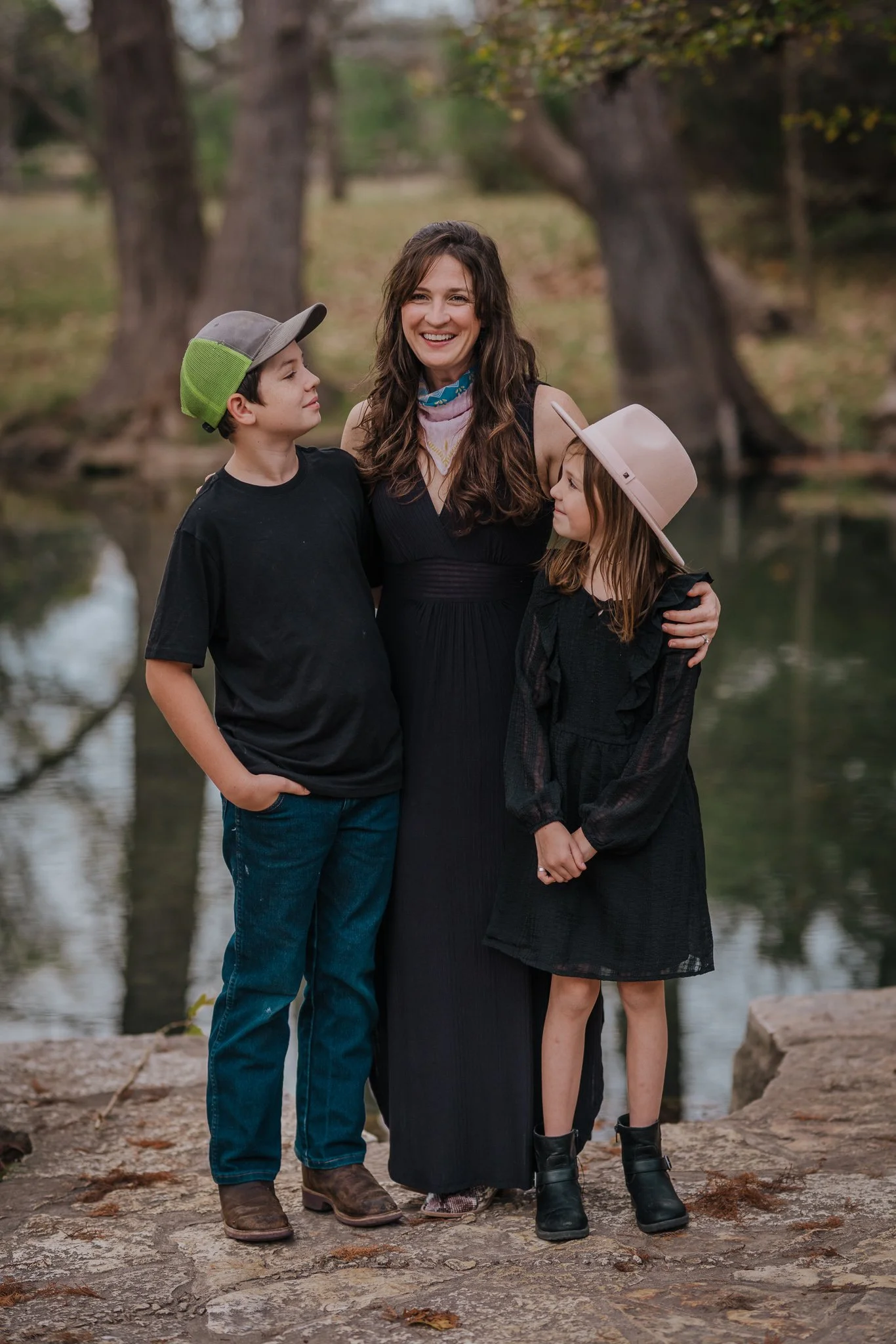 A woman with two children standing outdoors near a body of water in a park or forested area during fall, smiling at the camera. The boy is wearing a black shirt and green cap, and the girl is wearing a black dress and pink hat.