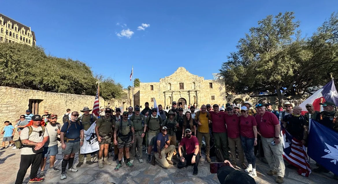 Group of people outdoors at a historical site, some holding American and Texas flags, with ancient stone buildings and trees in the background.