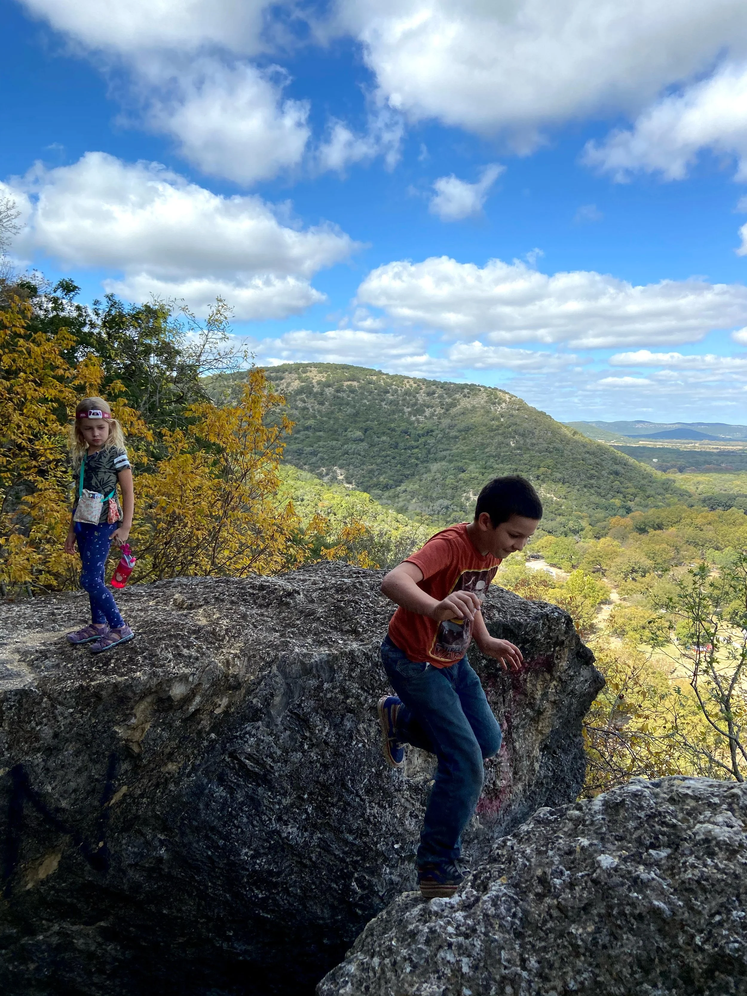 Two children, a girl and a boy, are climbing on large rocks outdoors with a scenic landscape of trees, hills, and a partly cloudy sky in the background.