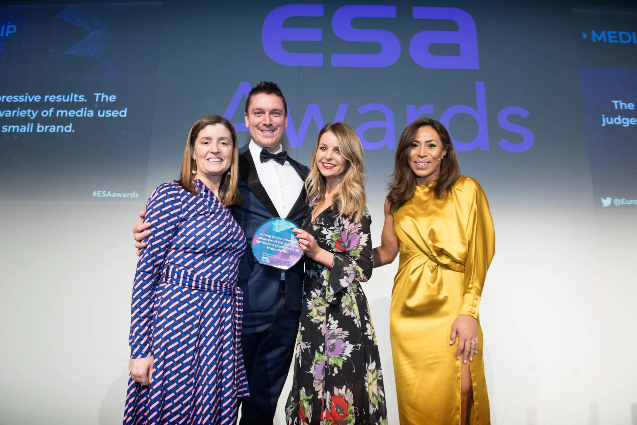 Four people dressed in formal attire standing together on stage at an award ceremony. The man in the middle is holding an award plaque. Behind them is a large screen displaying the words 'ESA Awards' and social media handles.