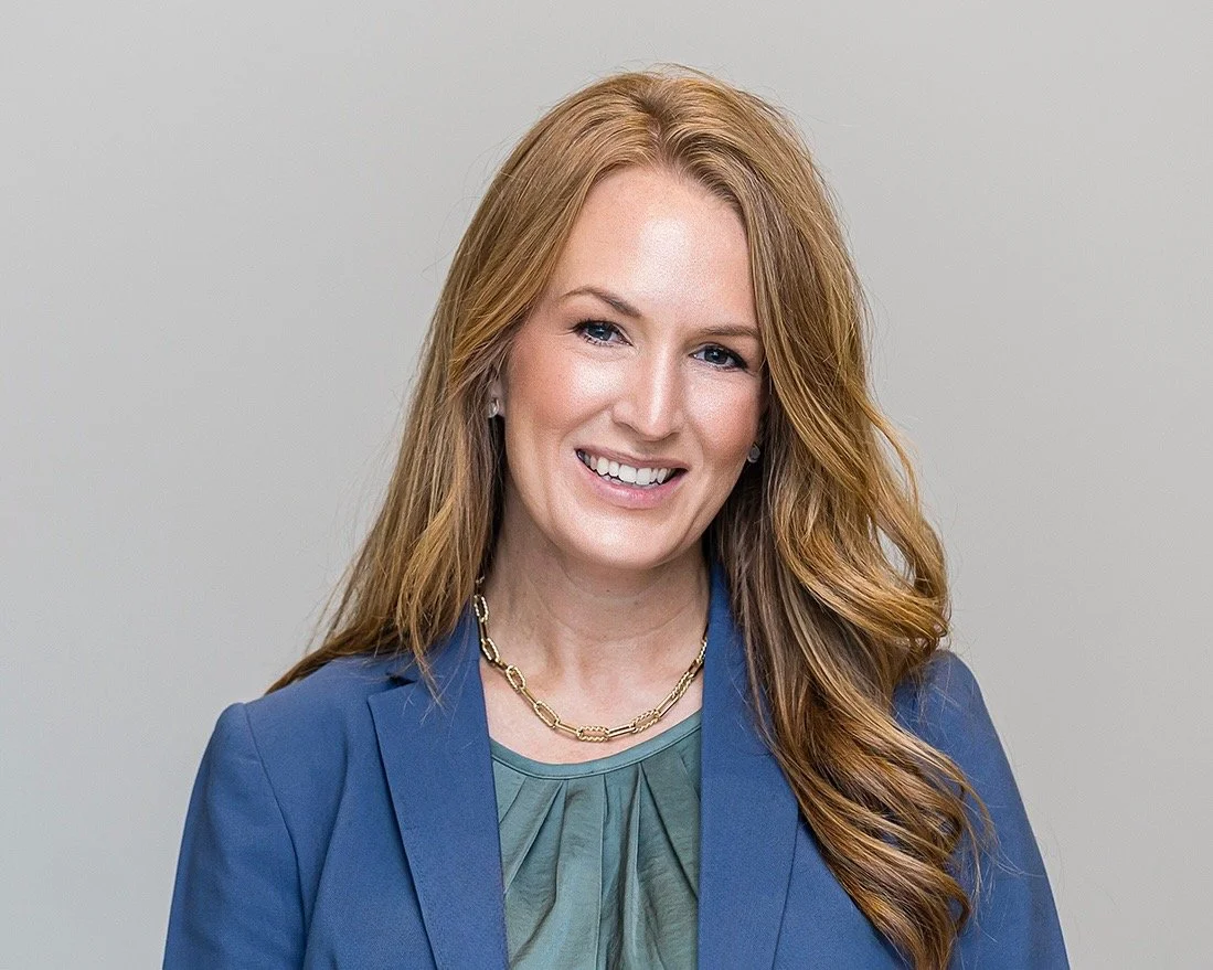 A woman with long red hair wearing a green blouse and blue blazer, smiling against a plain light gray background.