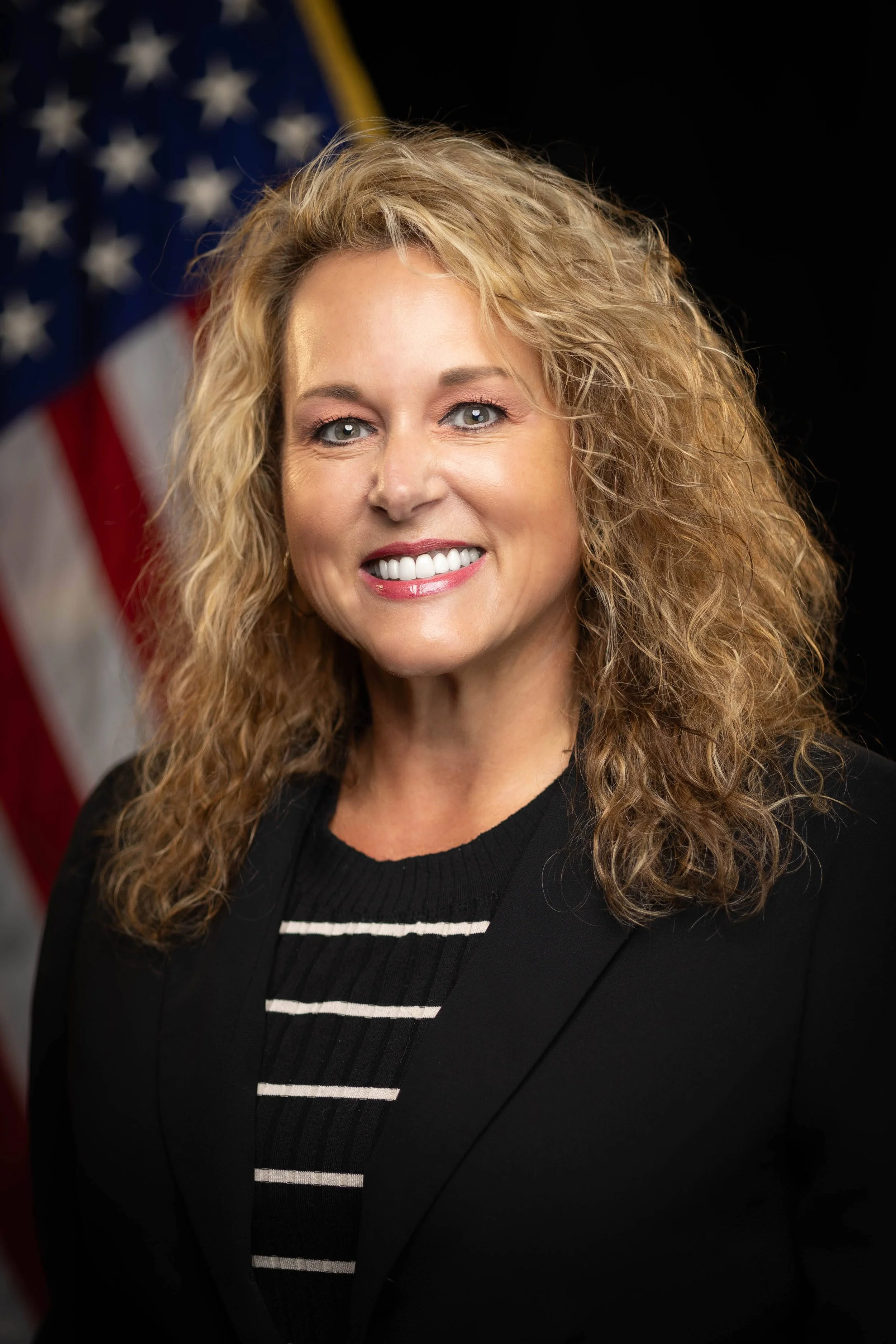 Portrait of a smiling woman with curly blonde hair, wearing a black blazer and a black-and-white striped top, with an American flag in the background.