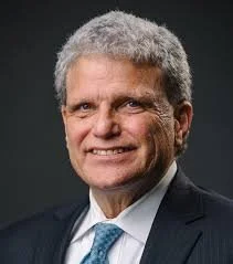 A professional portrait of a smiling man with gray hair wearing a dark suit and light blue tie against a dark background.