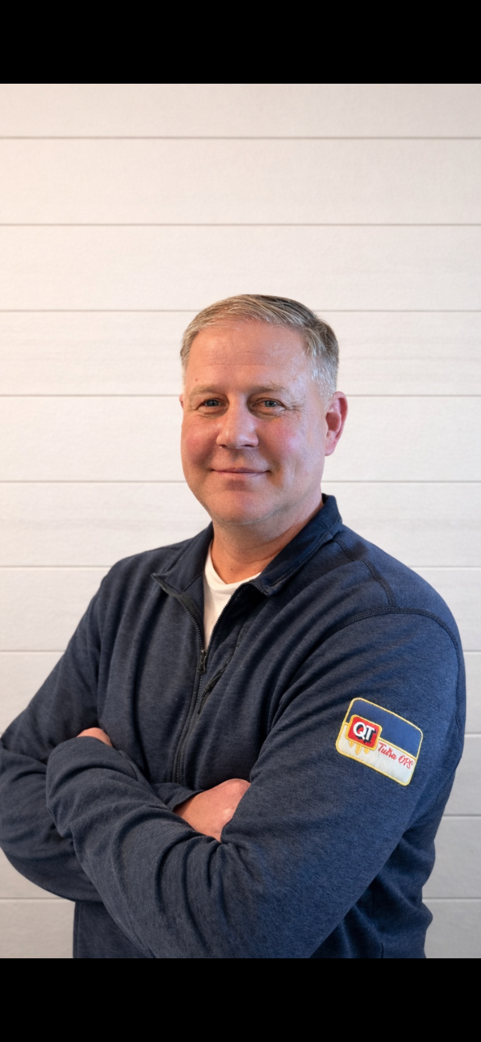 A smiling man with short blonde hair standing against a white paneled wall, wearing a navy zip-up jacket with a logo patch on the sleeve.
