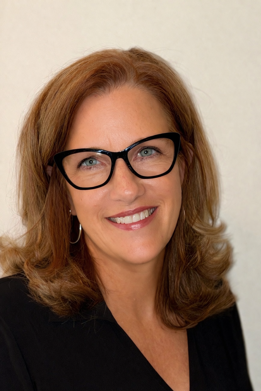 A woman with shoulder-length reddish-brown hair smiling, wearing purple sunglasses and a magenta top, outdoors with sunlight casting shadows on a concrete wall in the background.