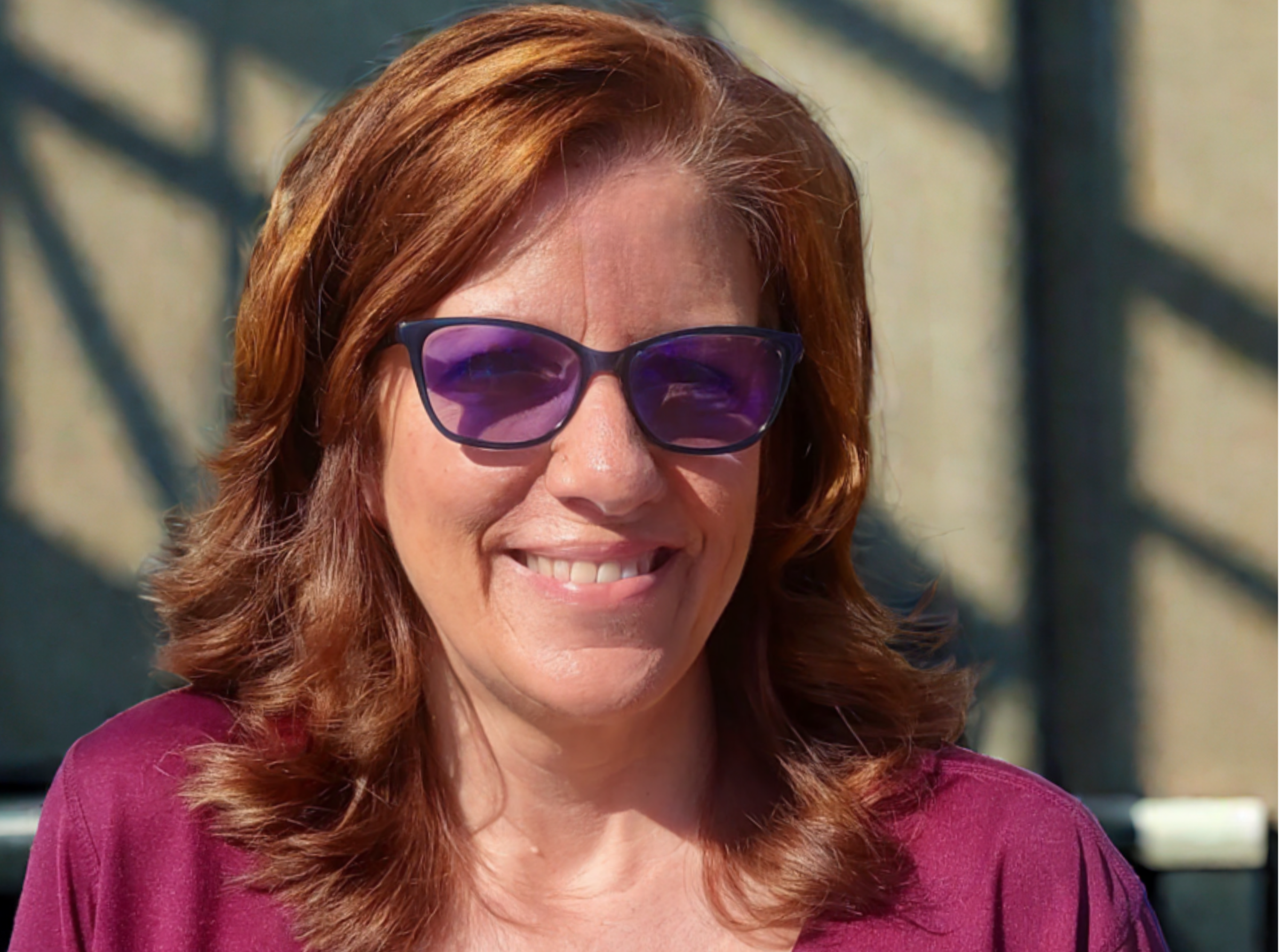A woman with shoulder-length reddish-brown hair smiling, wearing purple sunglasses and a magenta top, outdoors with sunlight casting shadows on a concrete wall in the background.
