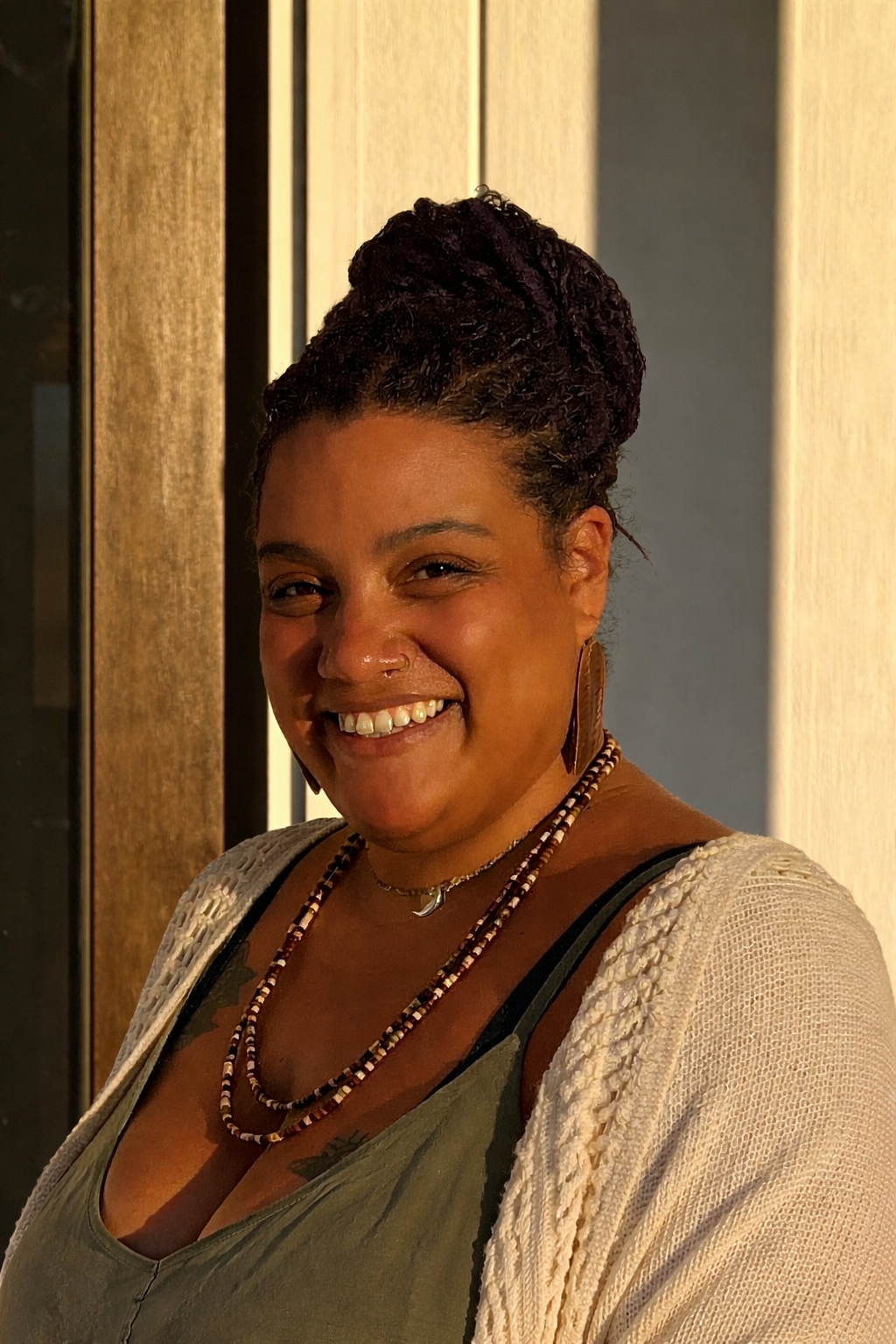 A woman smiling with hair styled in twists, wearing earrings, necklaces, a green top, and a light-colored cardigan, standing indoors near wooden panels.