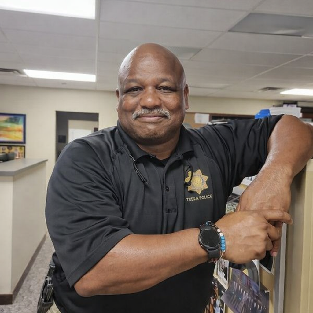 Man standing inside what appears to be an office environment. He is leaning casually against a cubicle partition with his arms resting comfortably. He is wearing a black polo shirt with a Tulsa Police badge emblem on the chest.