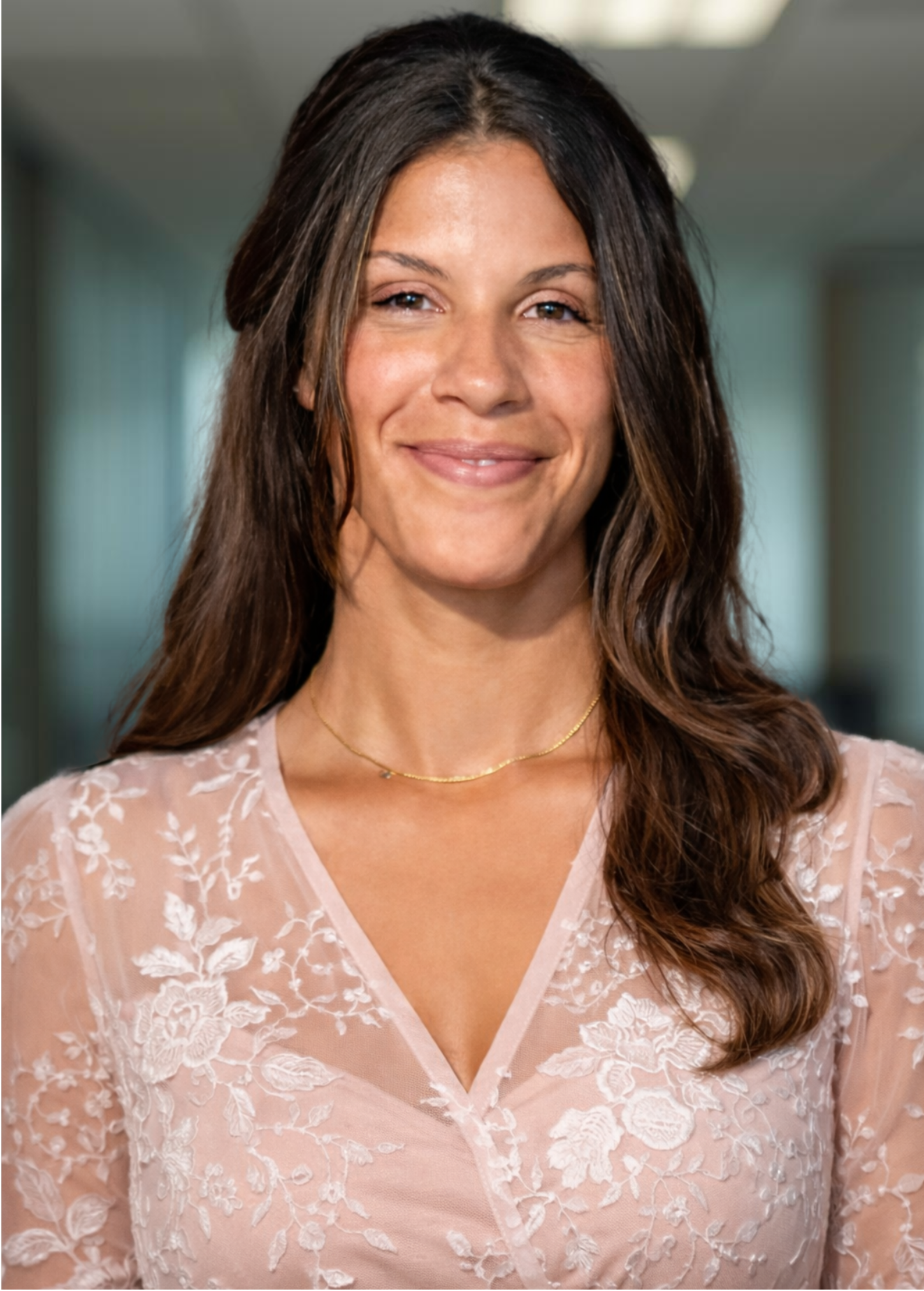 A woman with long brown hair smiling and wearing a pink floral lace top with a delicate gold necklace, indoors with a softly lit background.
