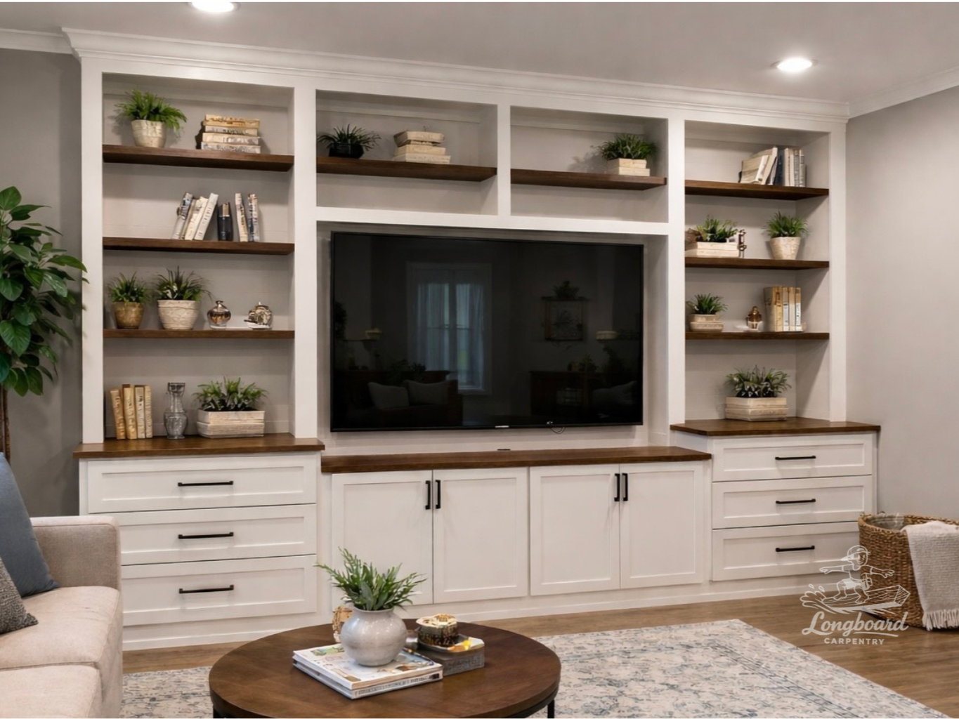 Living room entertainment center with a large flat-screen TV, surrounded by custom pained white cabinetry with brown wooden shelves by Longboard Carpentry, decorated with books and potted plants, and a coffee table.