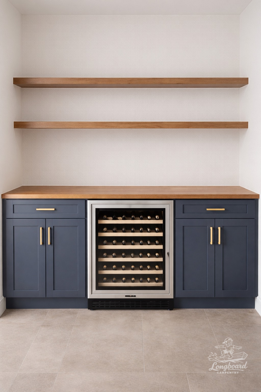A built-in wine cooler with a glass door, flanked by blue cabinets with gold handles, set beneath a wooden countertop and two wooden shelves against a white wall.