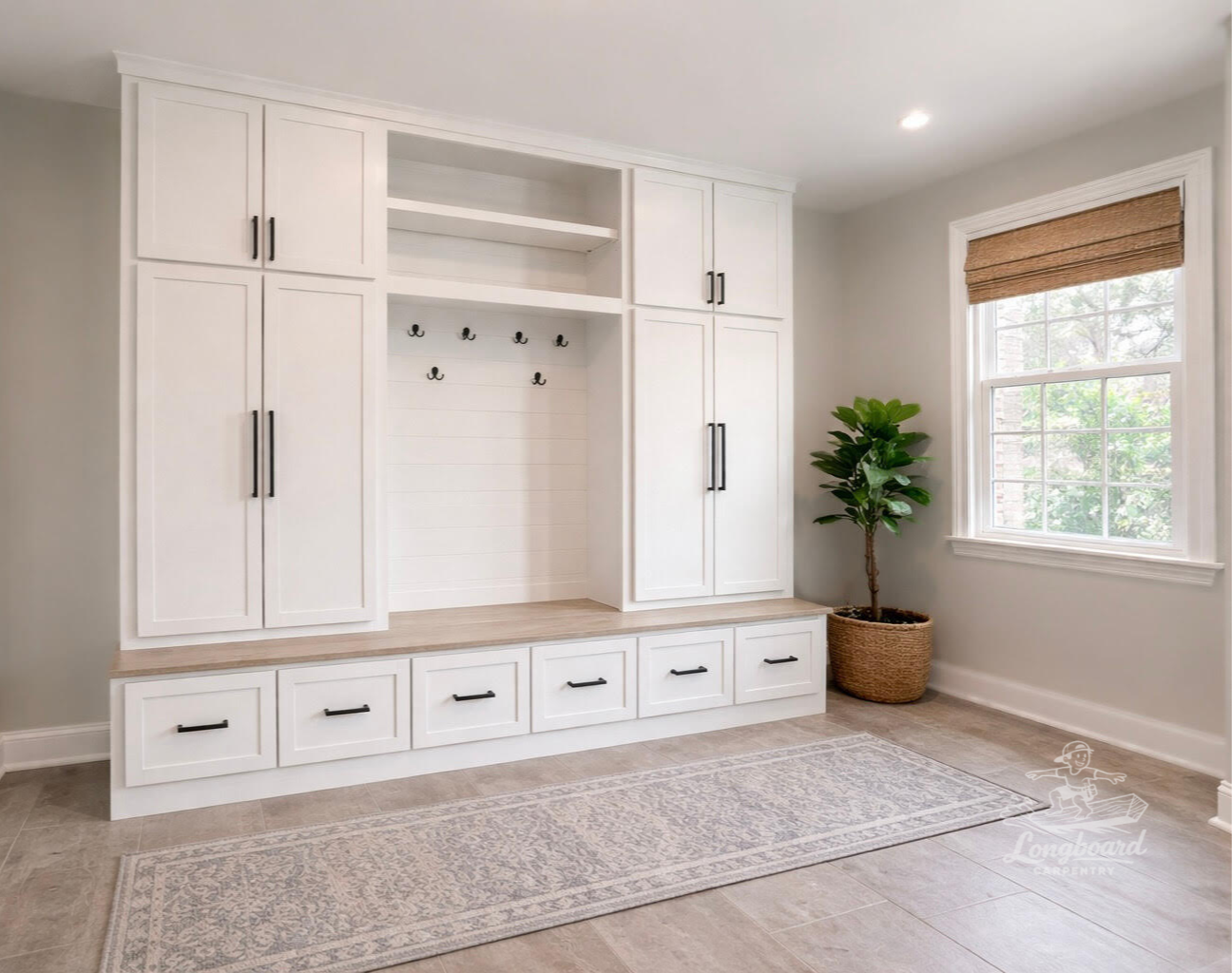 White custom built-in mud locker with cabinets, drawers, and hooks by Longboard Carpentry, next to a window with a bamboo shade, a potted plant, and a light-colored rug on tile flooring.