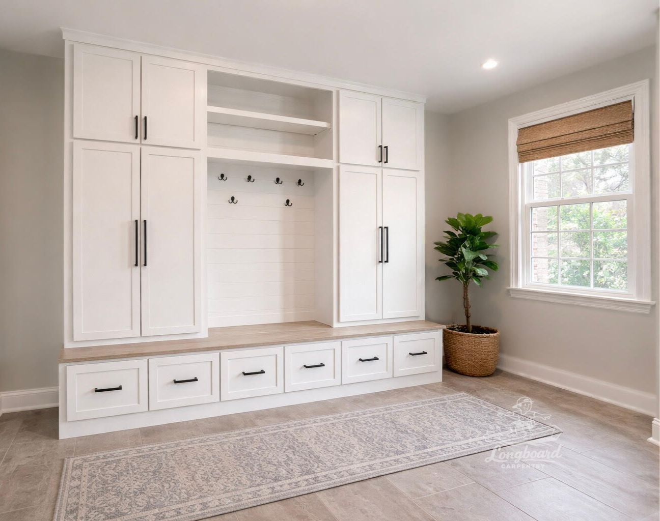 Bright contemporary mudroom with built-in white mud lockers with black handles, a wooden bench top, hooks, and open shelves. A large window with a brown woven shade, a potted green plant, and a patterned rug on tiled floor.
