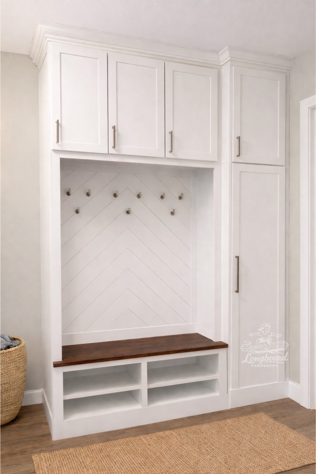 White built-in entryway mudroom mud locker with cabinets, hooks, a bench with a dark wooden top, cubby storage, and crown molding by Longboard Carpentry of Illinois. There is a woven basket on the left side and a natural fiber rug on the floor.