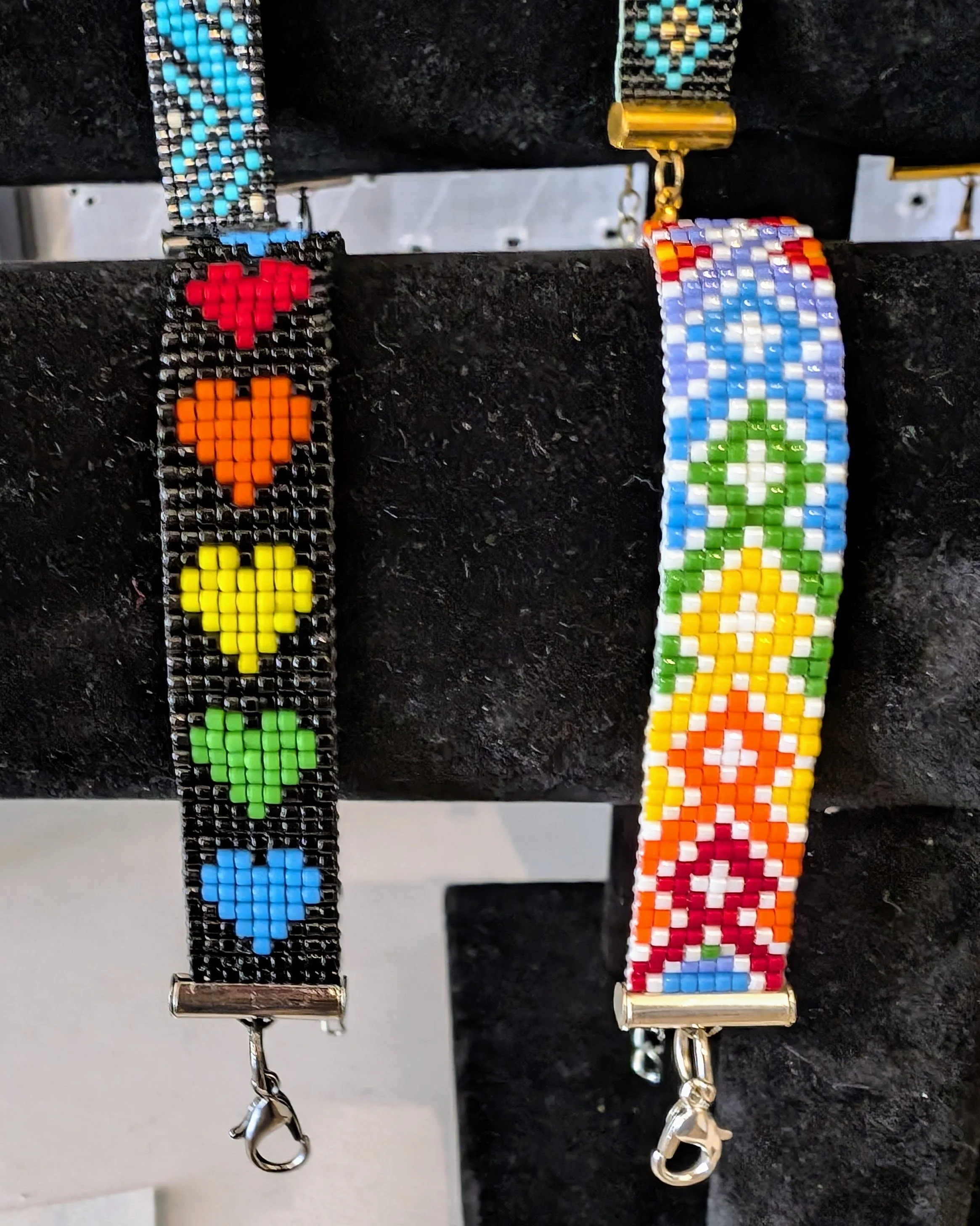 Close-up of two beaded keychains on a black surface, one featuring a red heart, a yellow flower, and a green leaf, the other with a rainbow pattern and a peace sign.