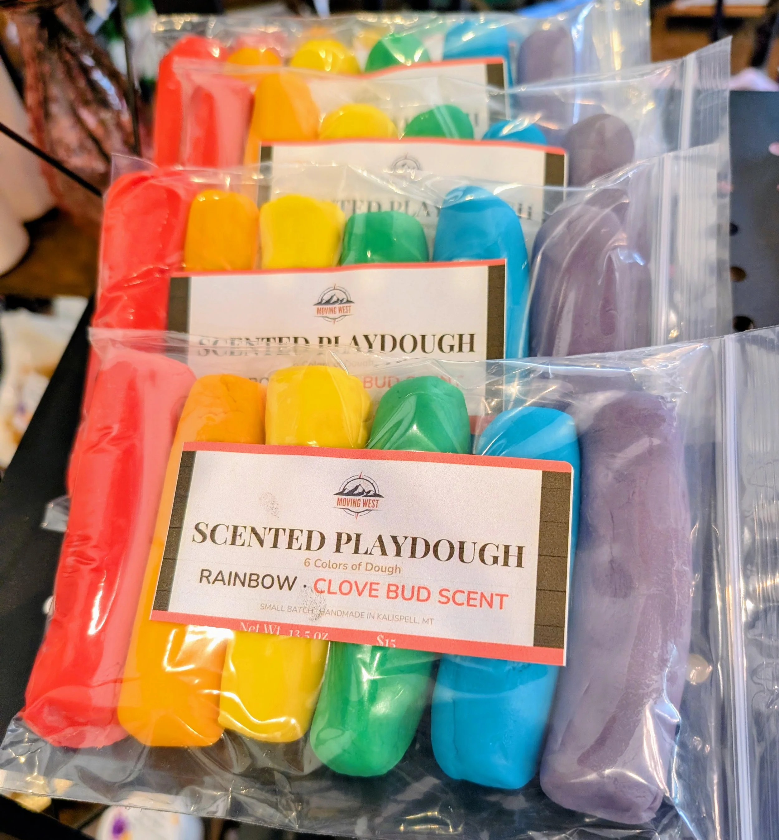 Assorted colored playdough packets with labels indicating scents like rainbow, clove bud, and lavender, in clear plastic packaging.