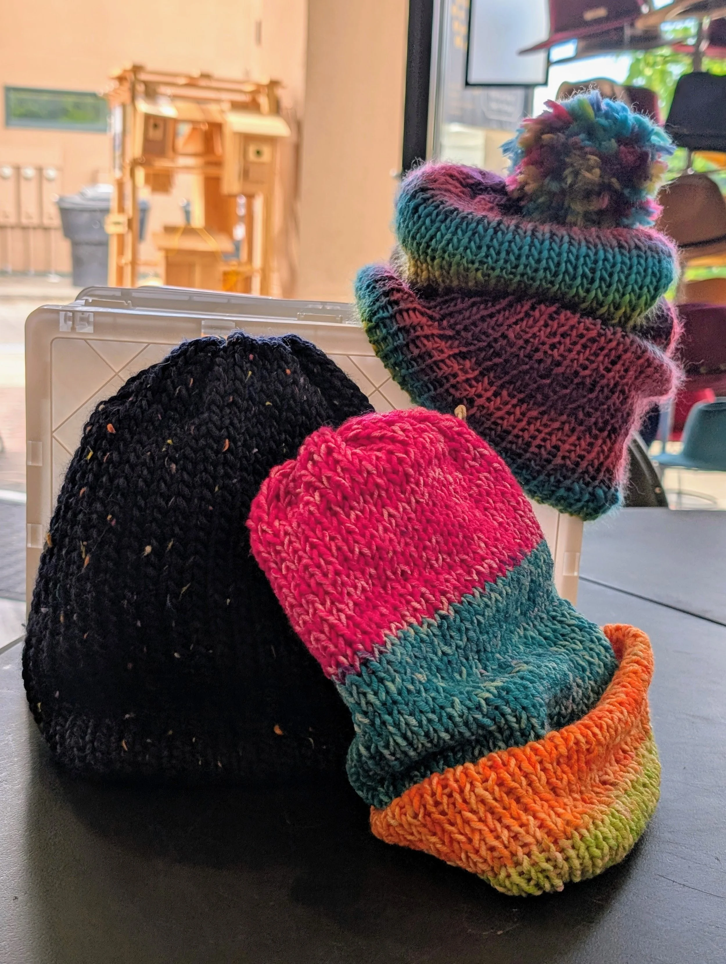 Colorful knitted beanies with pom-poms on a table, with wooden furniture and a television in the background.