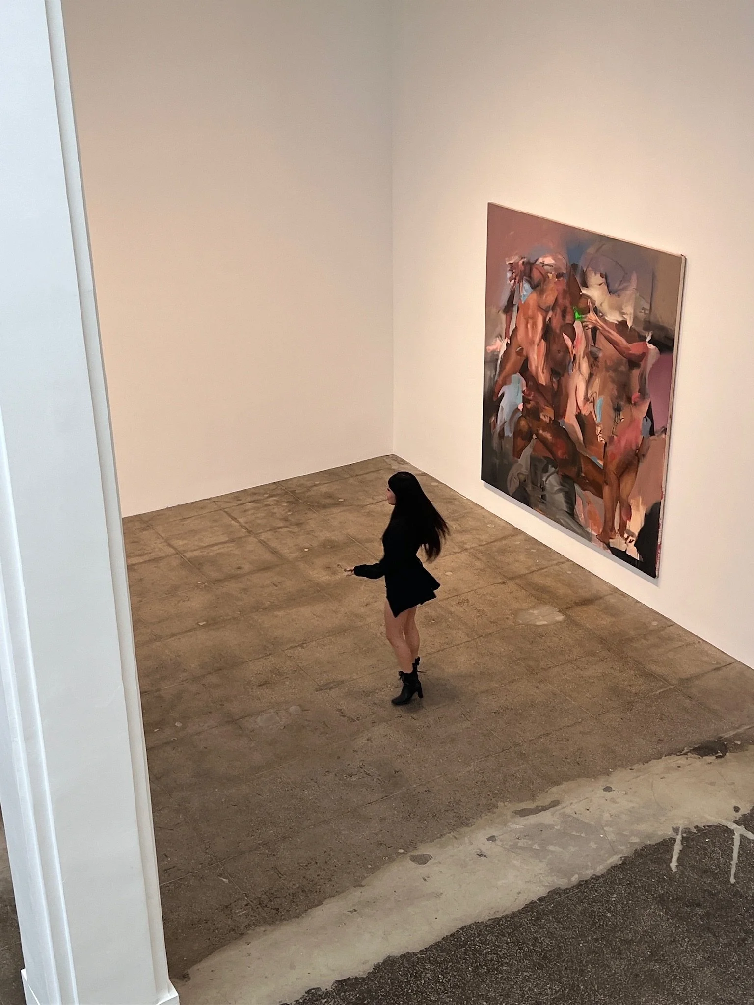 A woman in a black dress and high-heeled boots standing in an art gallery, viewing a large abstract painting of human figures on the wall.