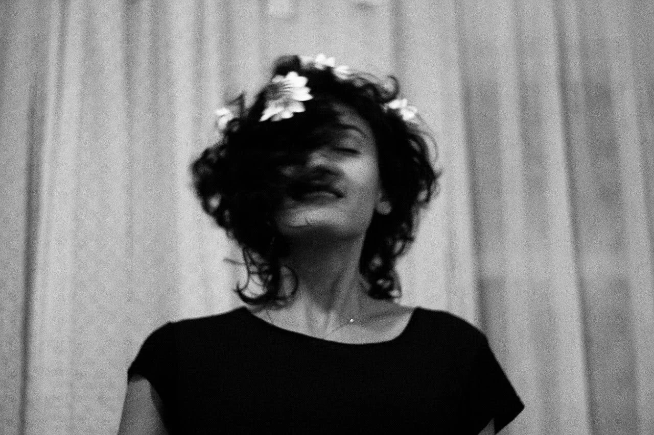 Mulher com cabelo encaracolado e florais, sorrindo com cabelo ao vento, usando camiseta preta, fundo de cortina