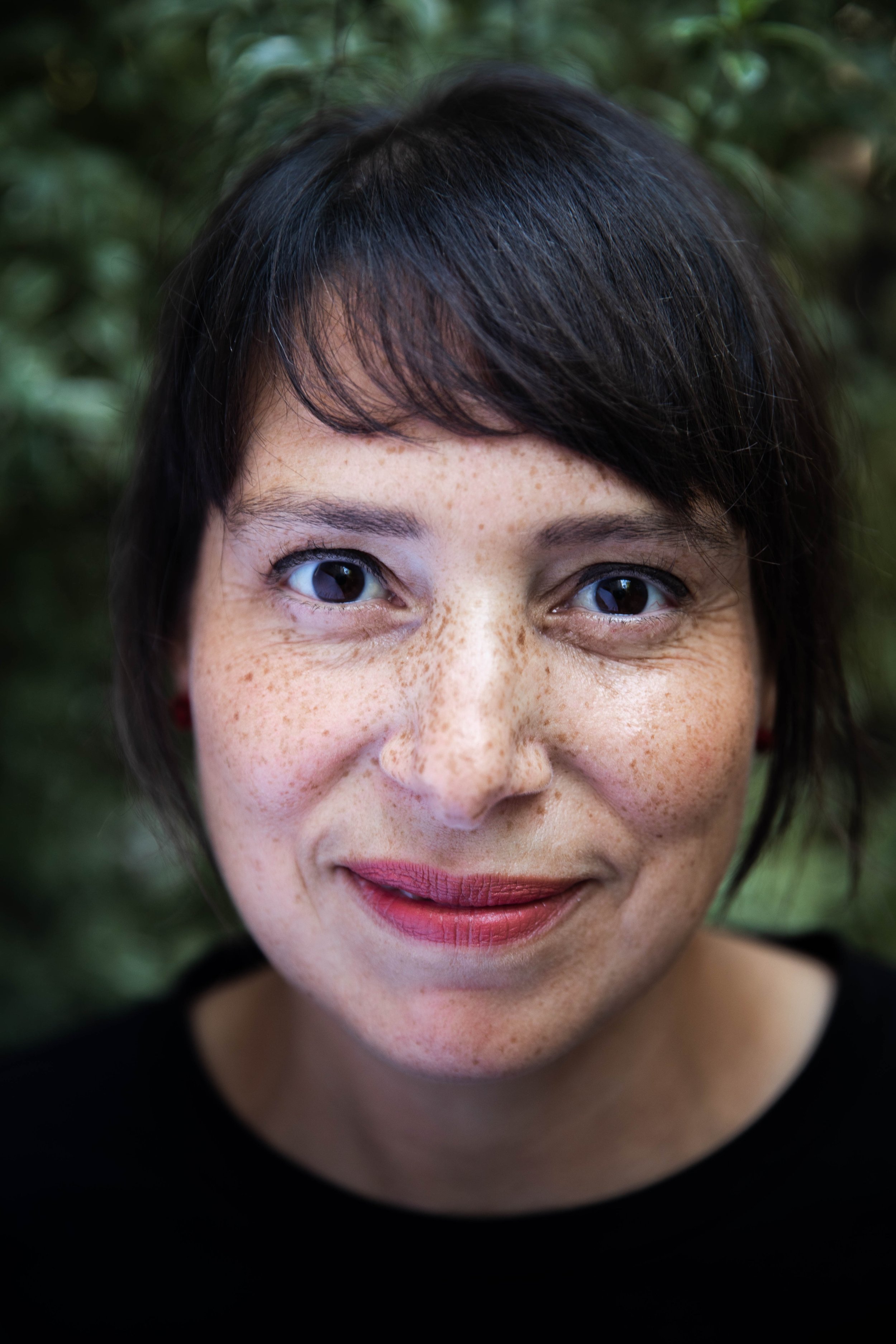 Close-up portrait of a woman with short dark hair, blue eyes, freckles, and wearing a black top, outdoors with green foliage in the background.