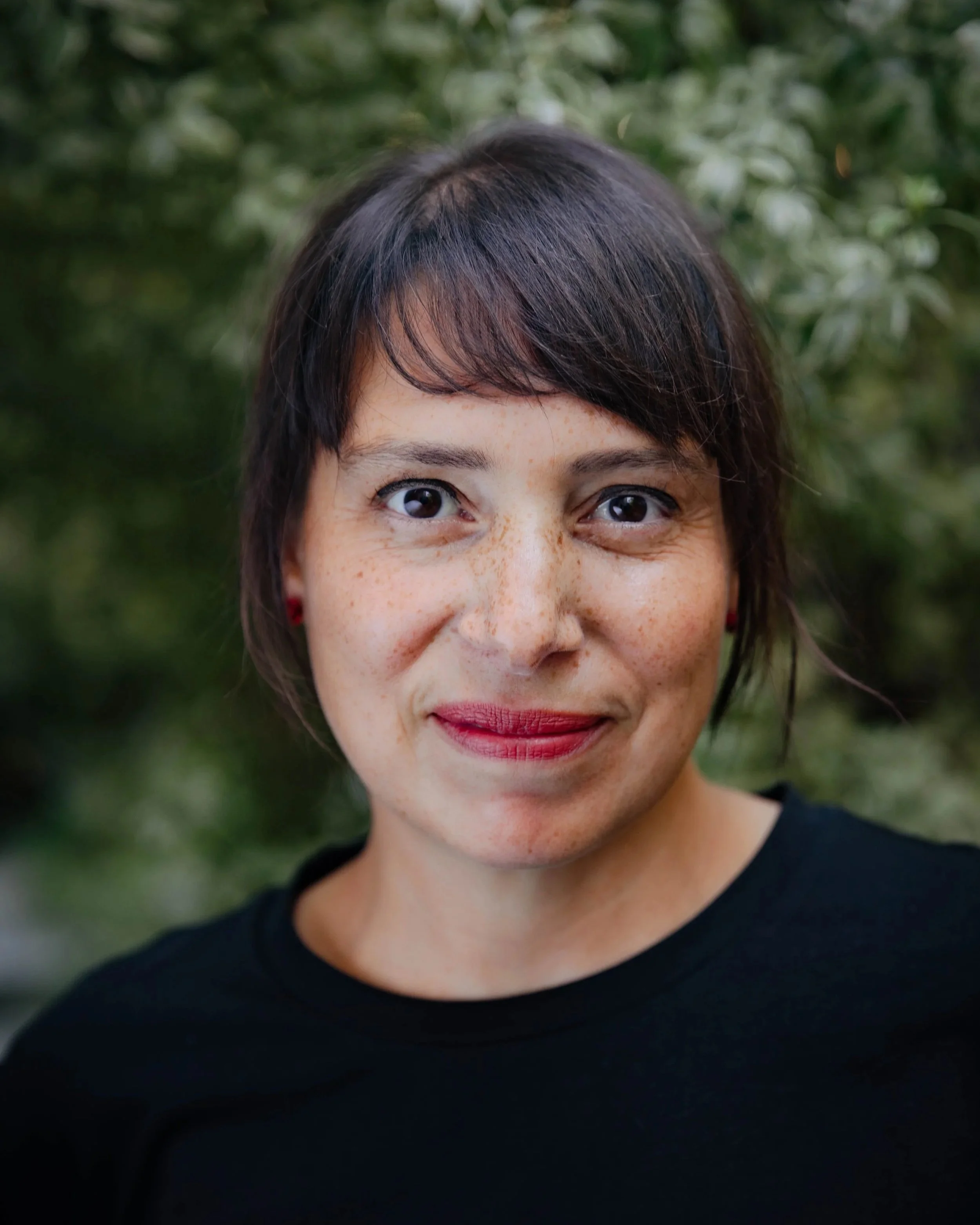 A portrait of a woman outdoors, with short dark hair, freckles, red lipstick, and a black top, standing in front of green foliage.