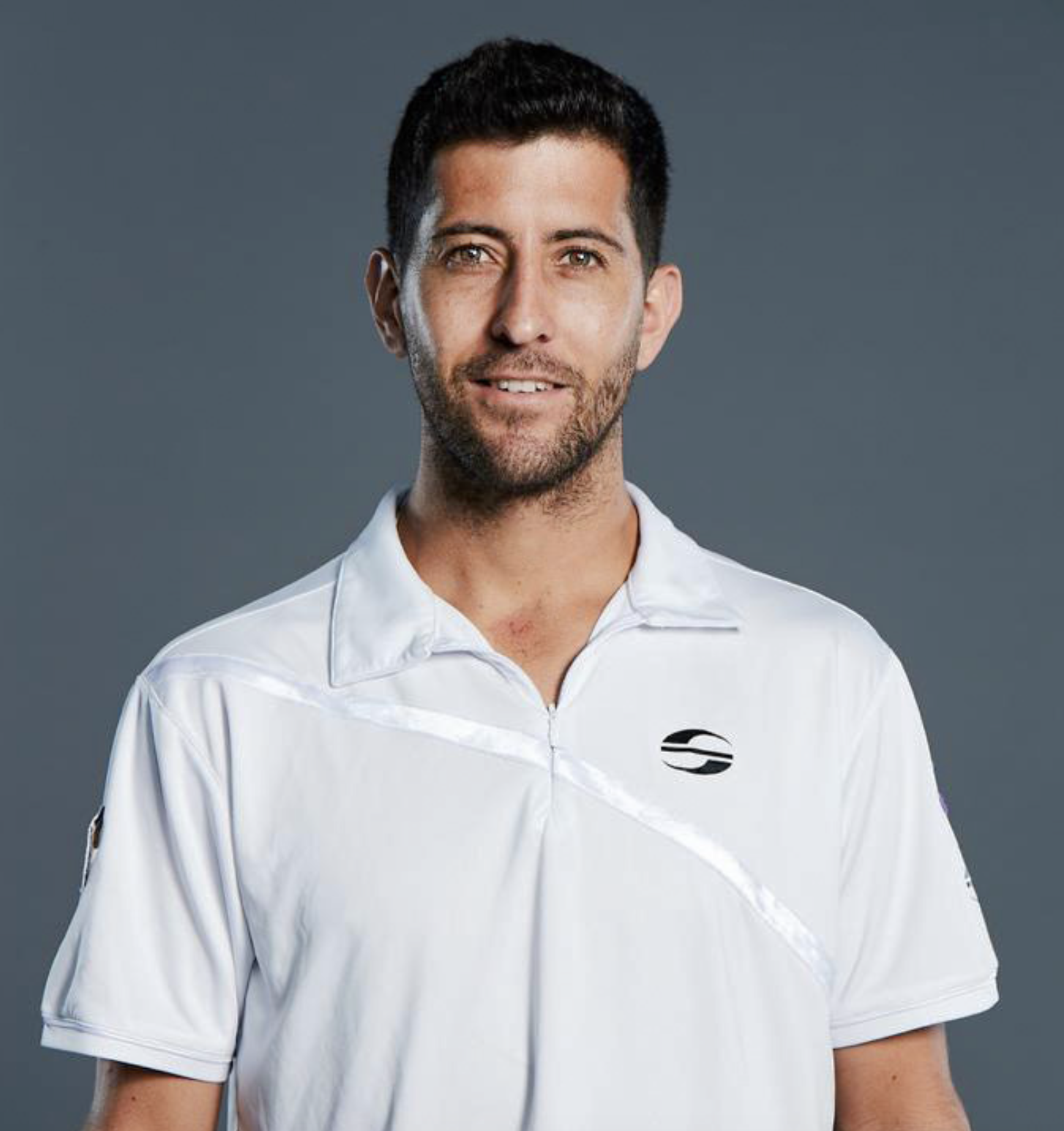A man with short dark hair and a beard, wearing a white polo shirt, standing against a dark grey background.