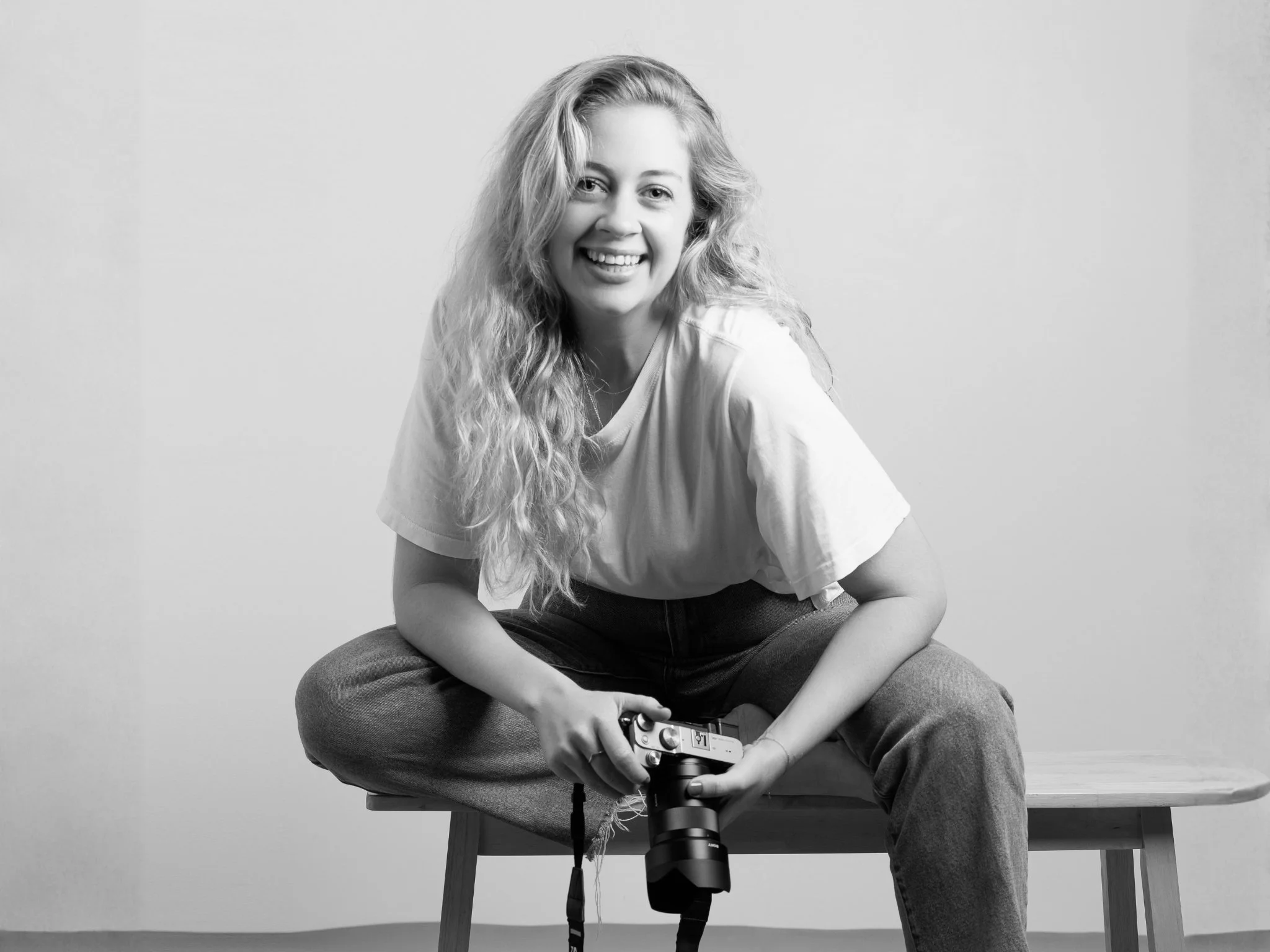 A young woman with long curly hair smiling while holding a camera, sitting on a wooden bench against a plain wall.