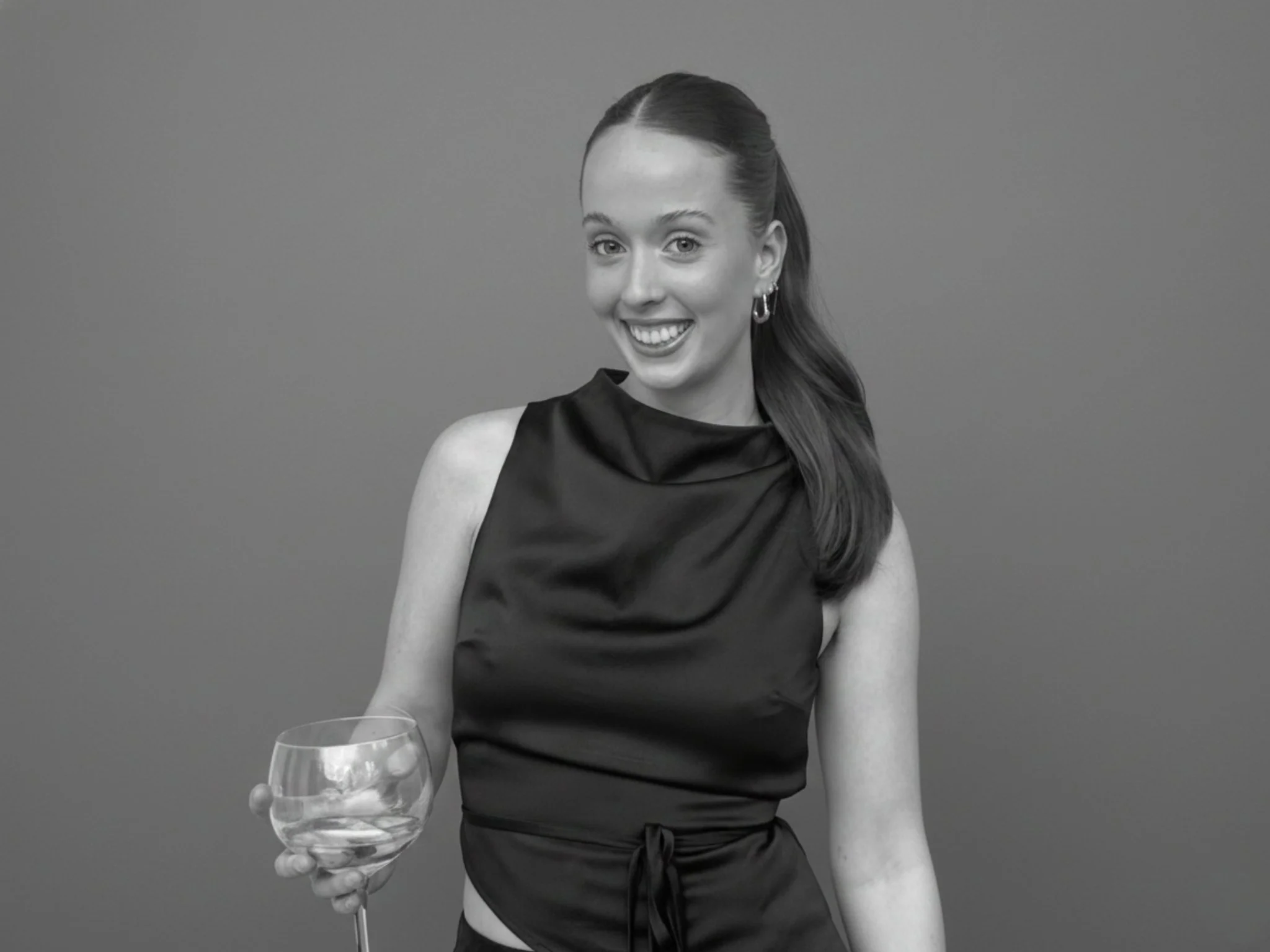 A young woman with long hair tied to one side, wearing a sleeveless black top, holding a wine glass, smiling, standing in front of a plain background.