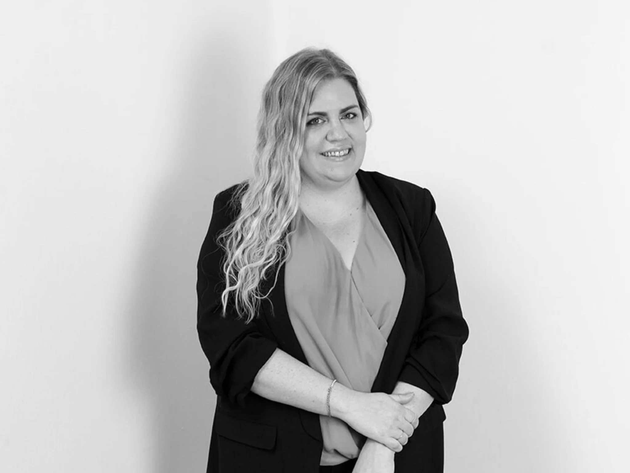 A woman with long, wavy hair wearing a blazer and blouse, smiling, standing against a plain wall.
