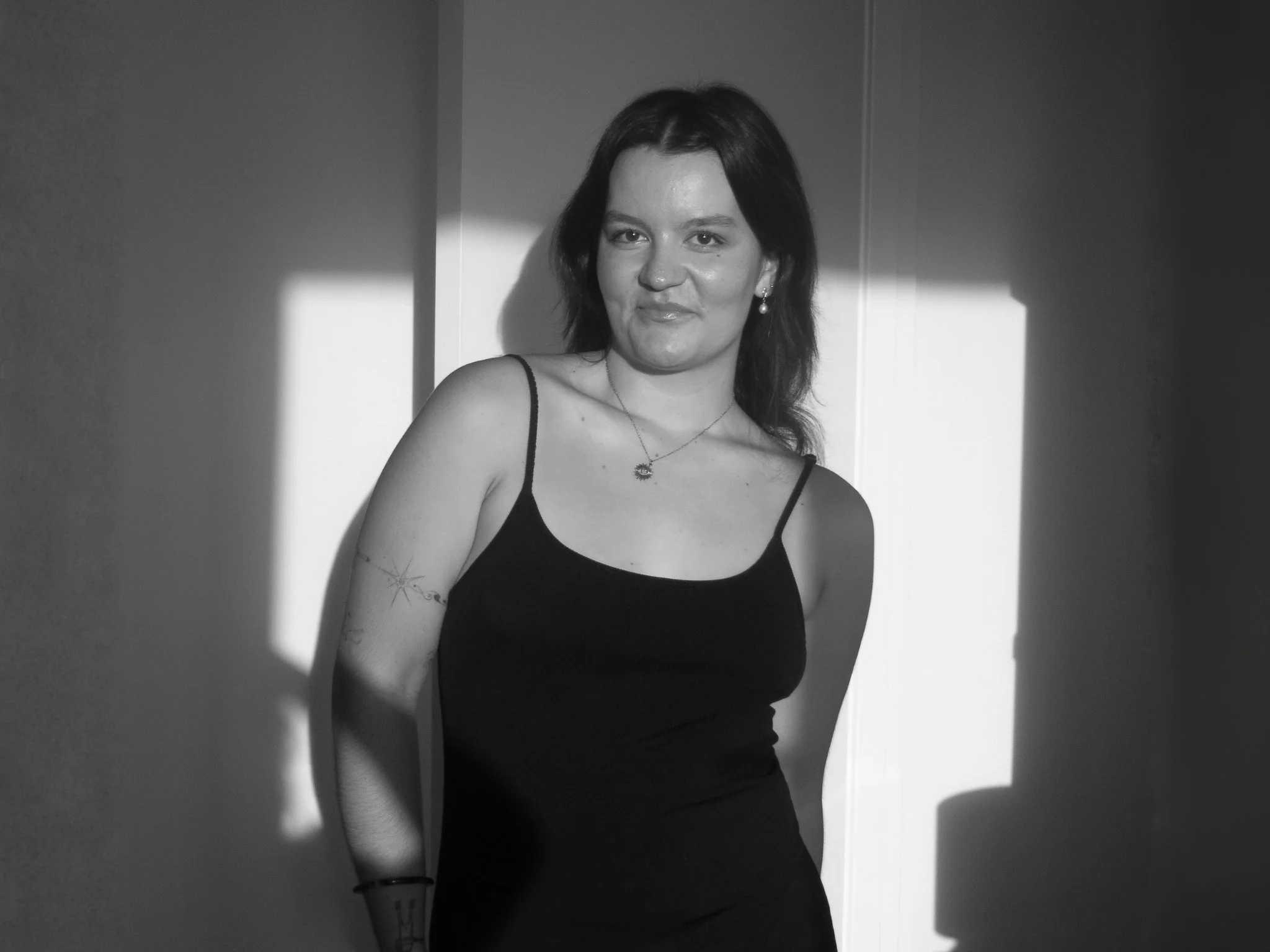 Black and white photo of a woman with dark hair in a black spaghetti strap dress, standing in front of a wall with light and shadow patterns.