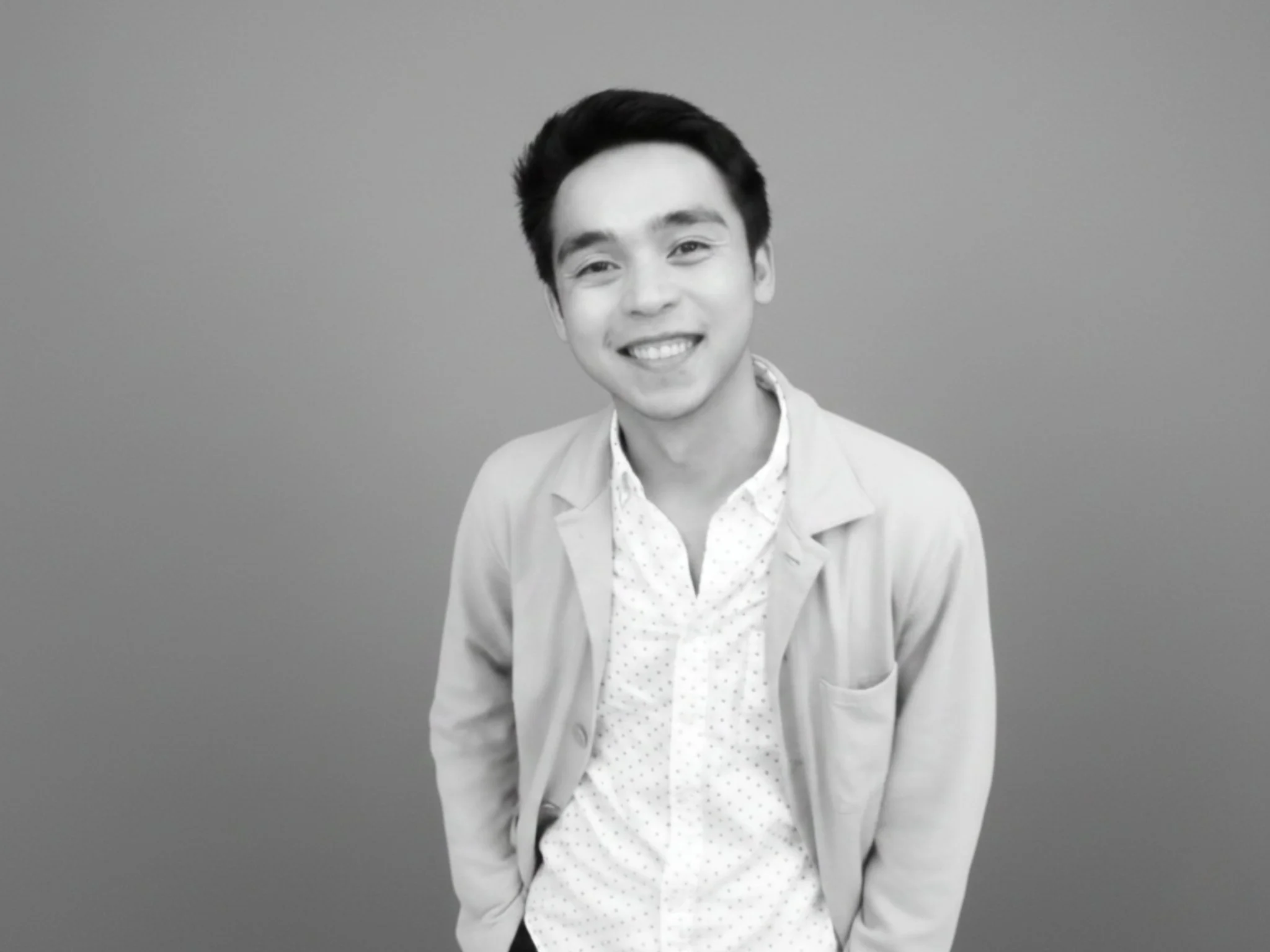 Black and white photo of a young man smiling and looking at the camera, wearing a light jacket over a collared shirt, standing against a plain background.